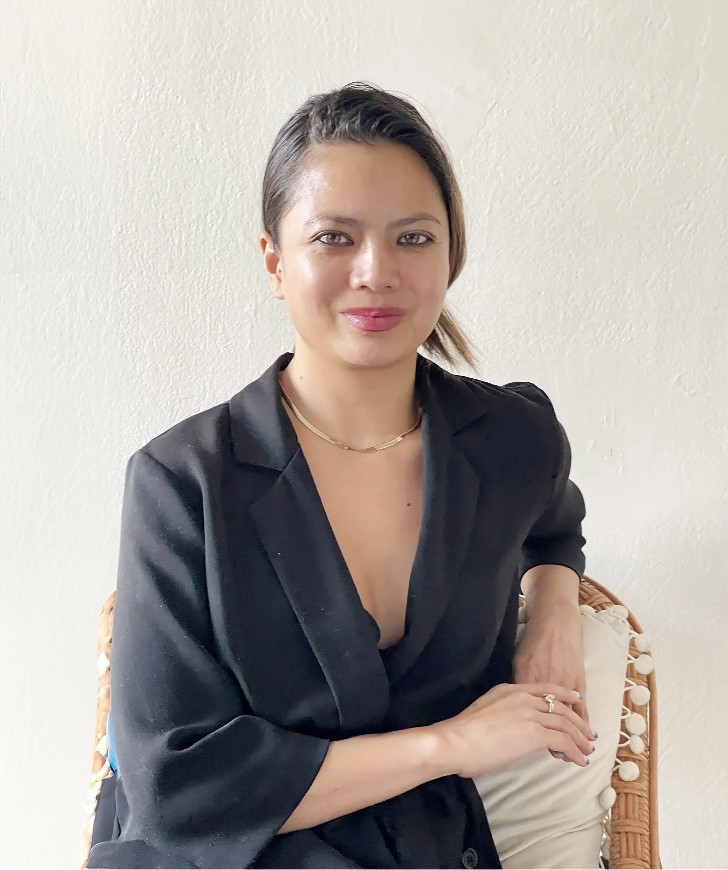 Person sitting in a chair wearing a black blazer with a gold necklace and rings, smiling at the camera.