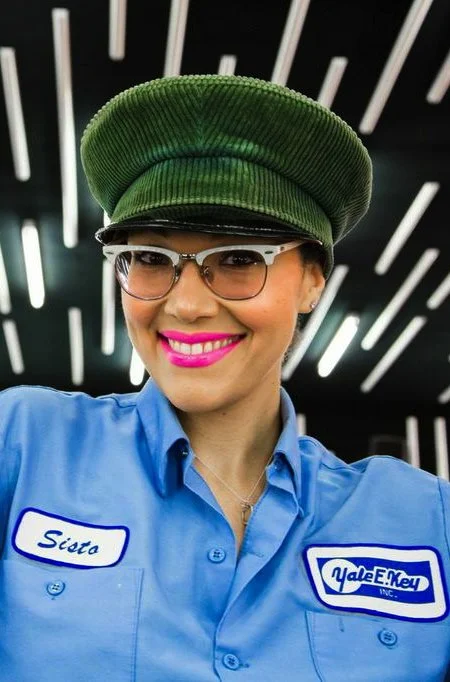 A woman wearing glasses, a green cap, and a blue work shirt with a name tag that says "Sista" and a Yale E. Kepp INC. patch, smiling against a background with vertical white and black lines.