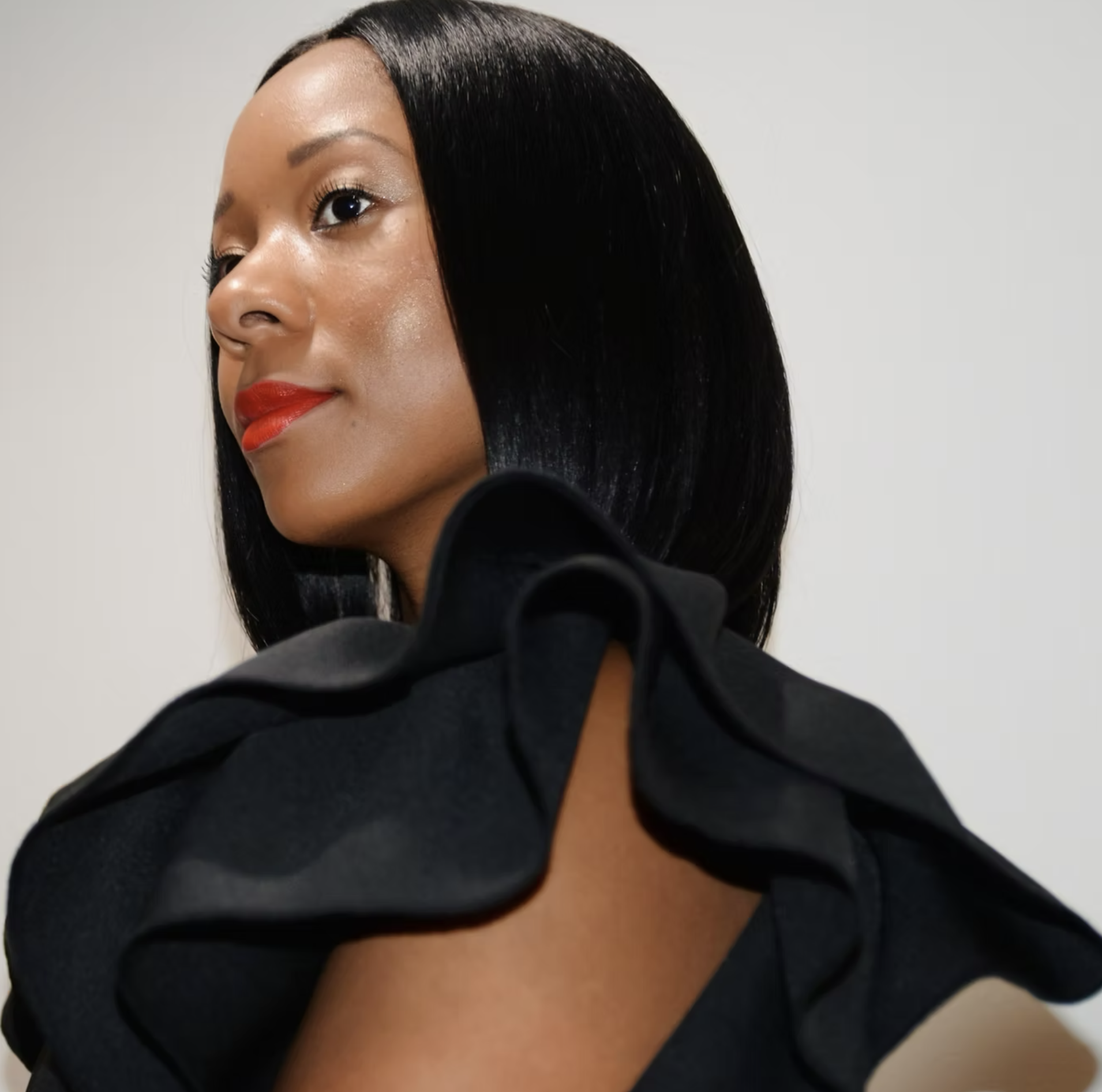 Close-up of an elegant woman with sleek black hair, red lipstick, and wearing a black outfit with large ruffled shoulder details, standing against a plain background.