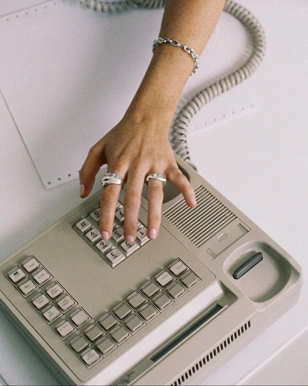 A hand with rings and a bracelet is pressing buttons on a vintage beige telephone with a numeric keypad and a coiled cord.