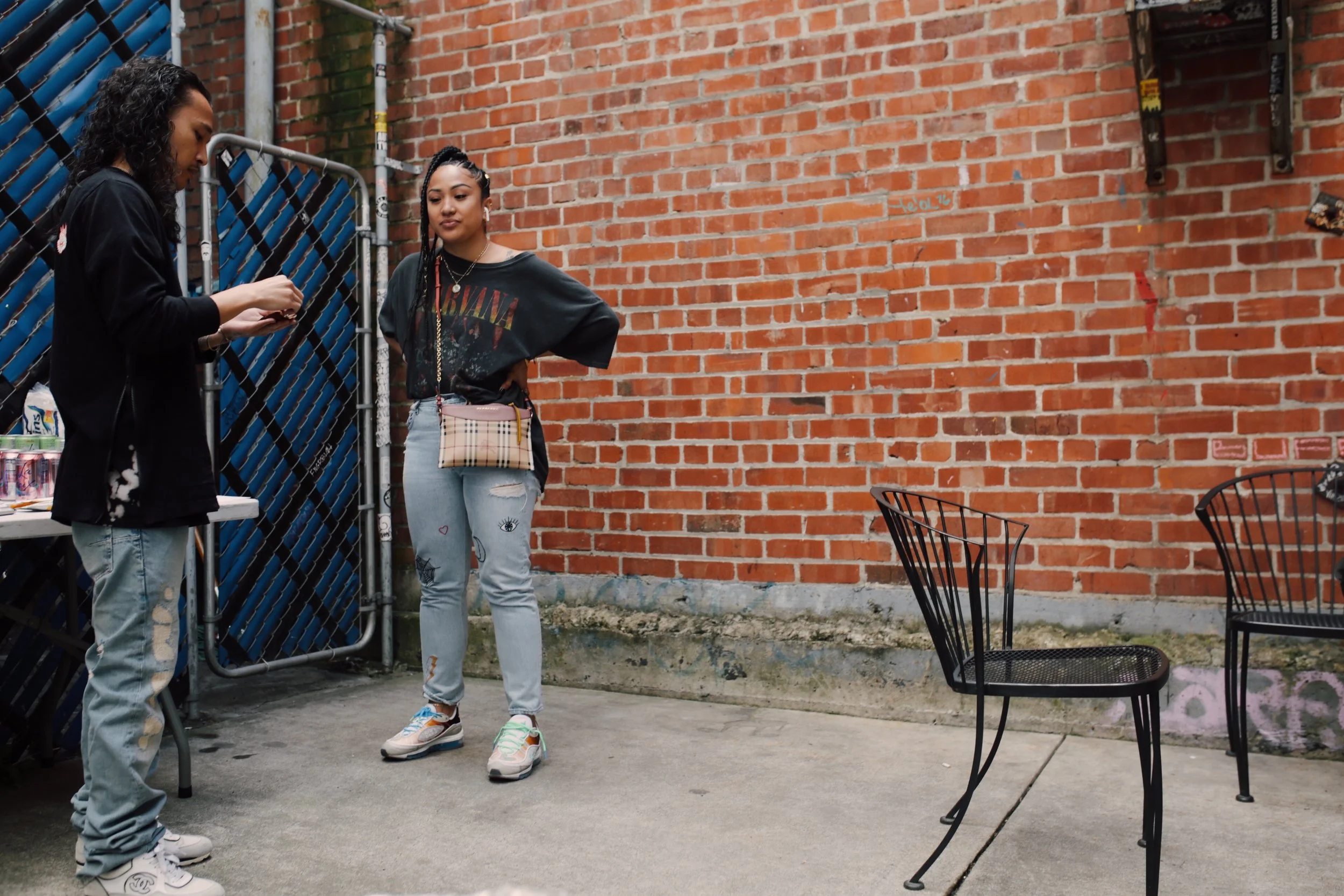 Two young women stand outside near a brick wall, one is looking at her phone while the other is looking at her, with a small beige purse across her shoulder. There are two black metal chairs and a table nearby.