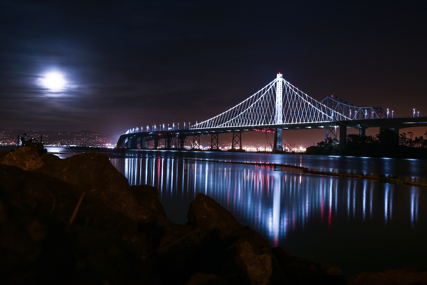 Night view of a lit-up bridge over water under a moonlit sky with city lights in the background and rocks in the foreground.