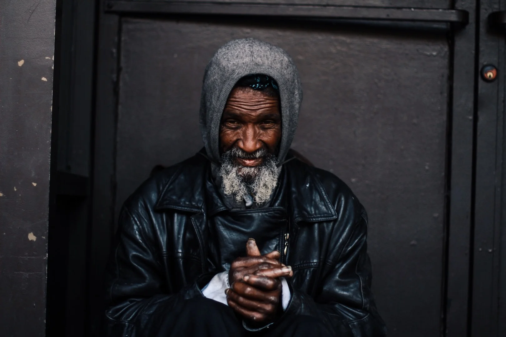An elderly man with a gray beard, wearing a gray beanie and a black leather jacket, sitting against a dark wall, looking at the camera with hands clasped.