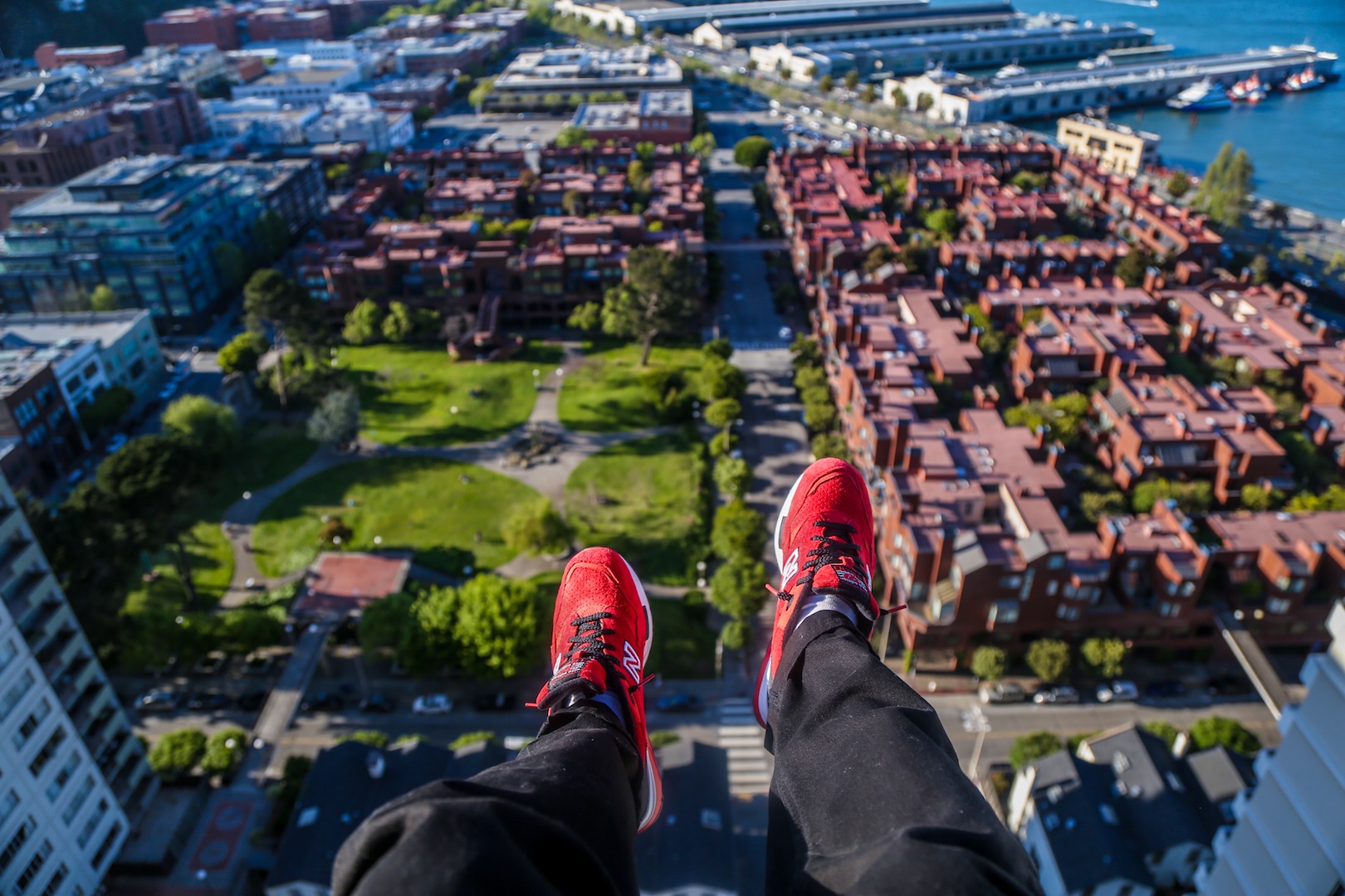 Person sitting on high rooftop ledge wearing black pants and red sneakers, overlooking urban cityscape with green park, residential and commercial buildings, and waterfront.