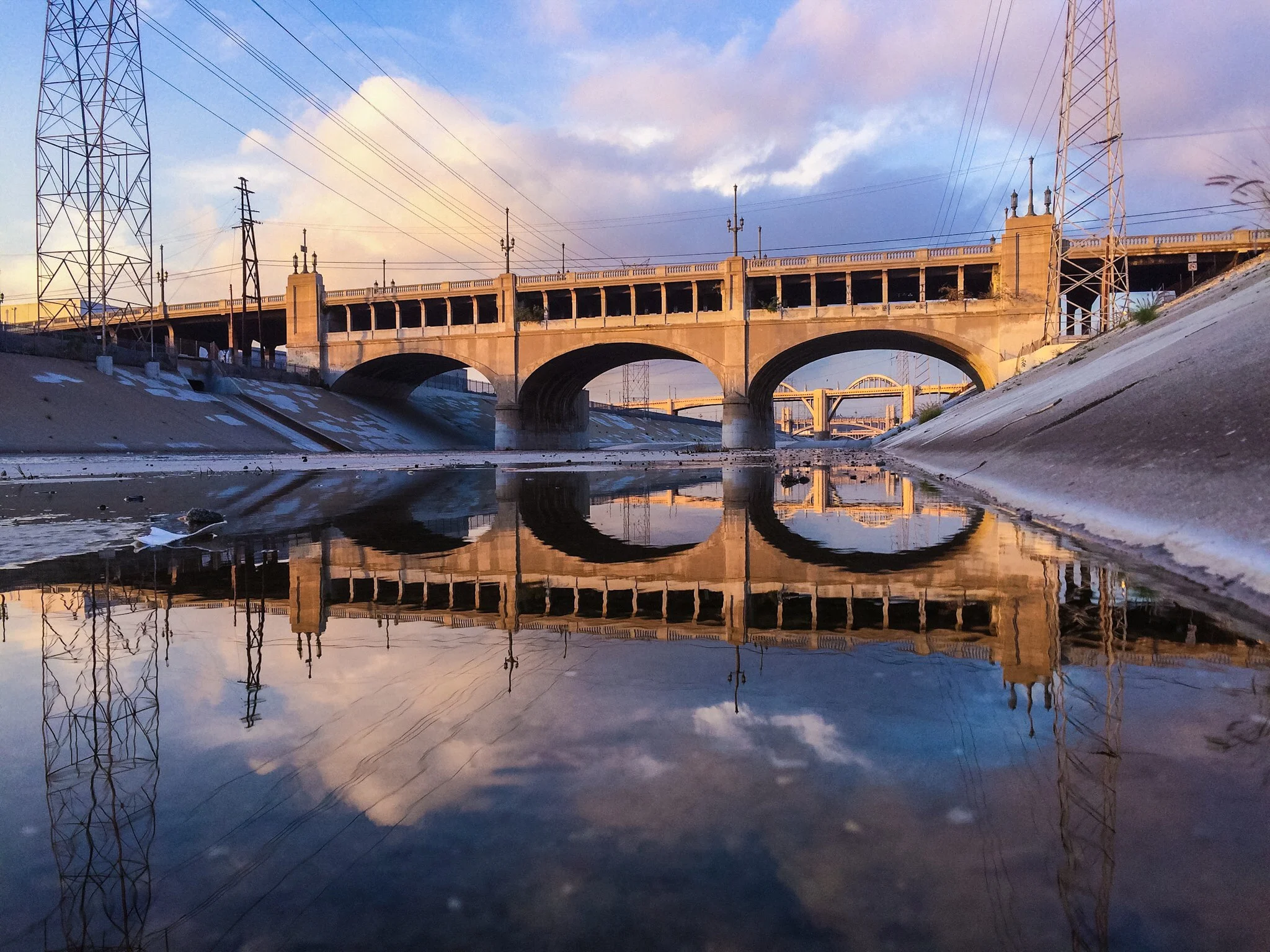 A bridge over a canal or river with its reflection visible in the water, at sunset or sunrise, with pylons and power lines in the scene.