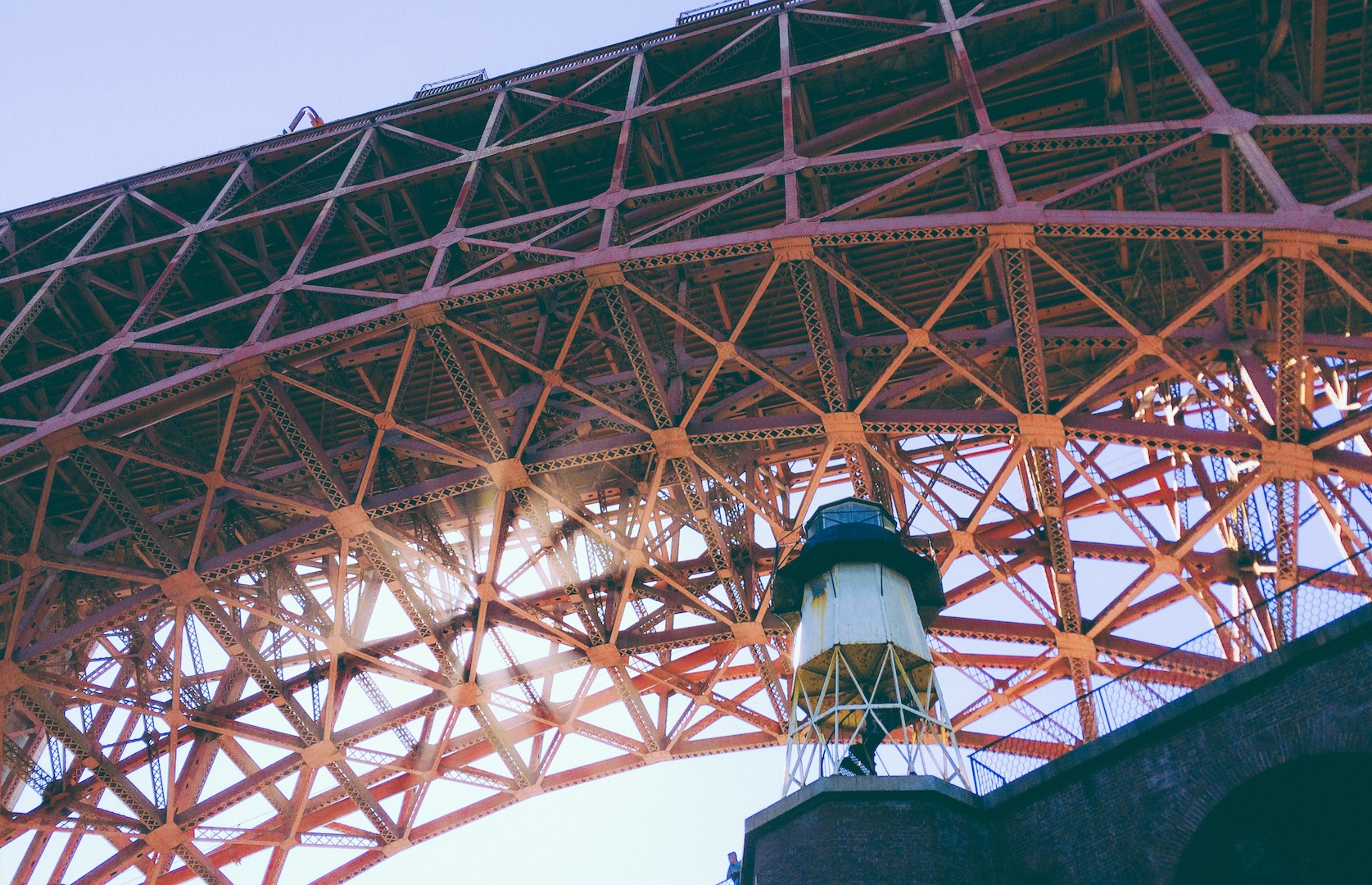 Close-up view of the underside of a large, red, industrial bridge structure, with a lighthouse-like light fixture on a platform beneath and sunlight shining through the trusses.