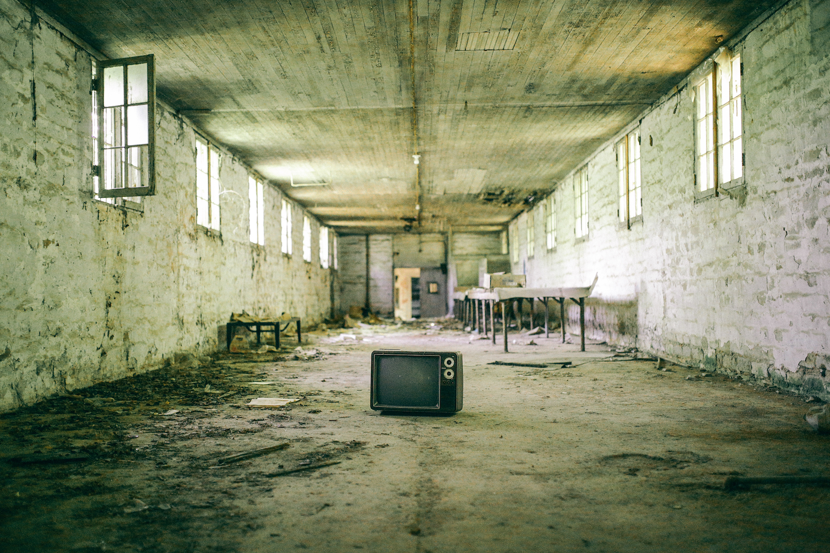 Abandoned room with broken windows, peeling paint, and an old television on the dusty floor.