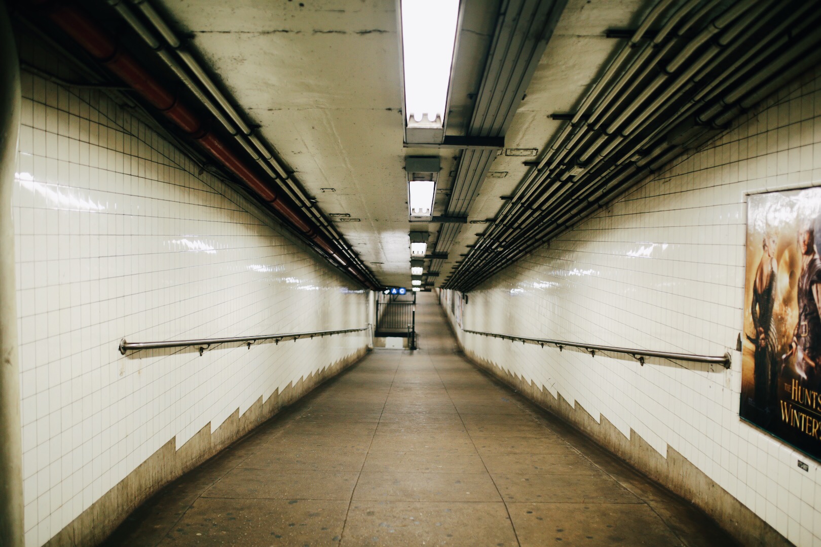 Underground pedestrian tunnel with white tiled walls, fluorescent lighting, and railings on both sides, leading to stairs at the end of the tunnel.