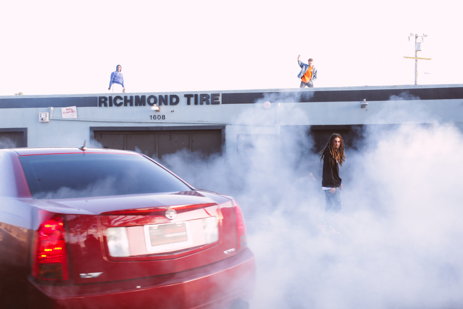 A red car is surrounded by white smoke in front of a building with a sign that says 'Richmond Tire.' Four individuals are on the building's roof, and one person is standing in the smoke, looking toward the camera.