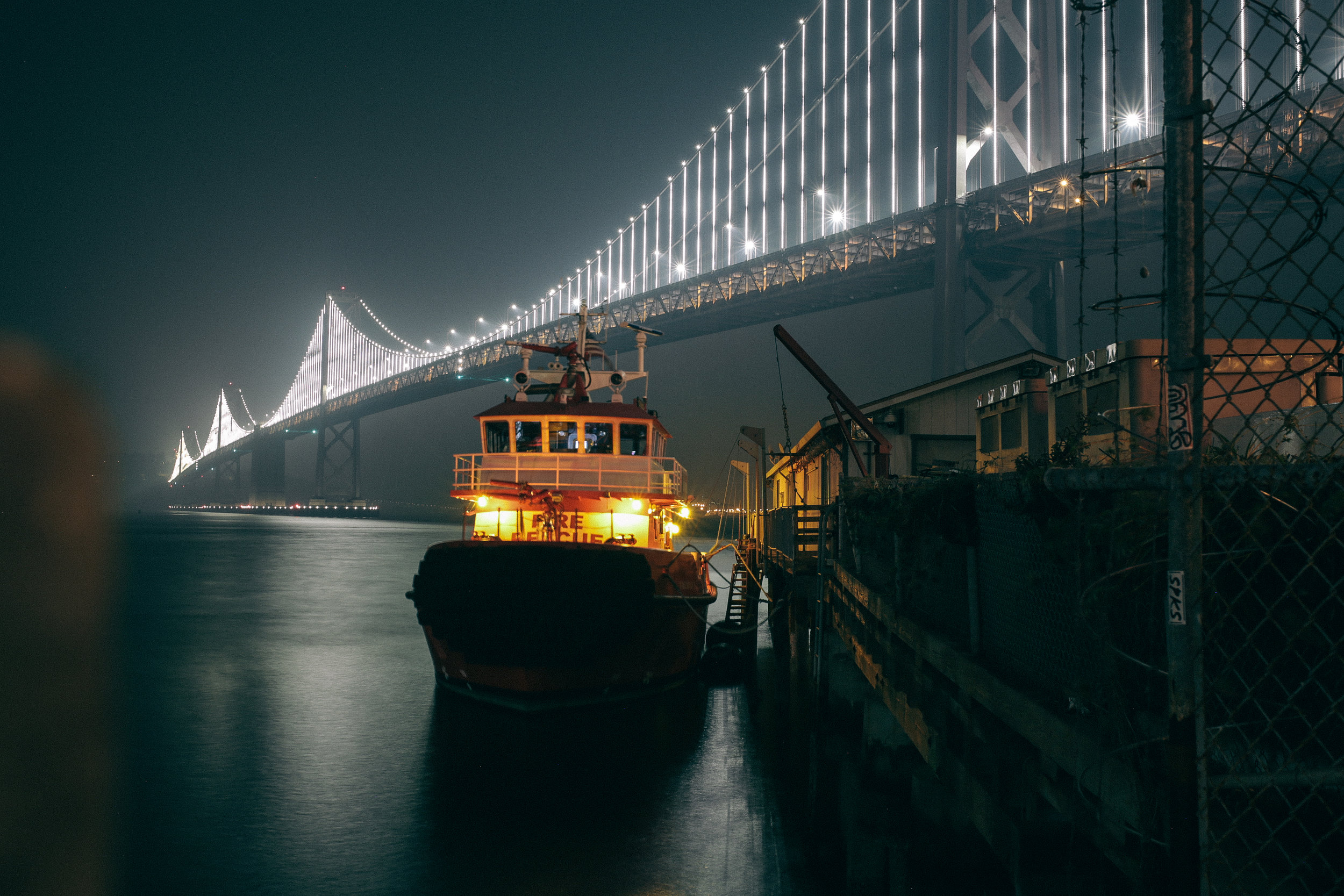 Nighttime view of a brightly lit tugboat docked at a pier with a large illuminated suspension bridge in the background.