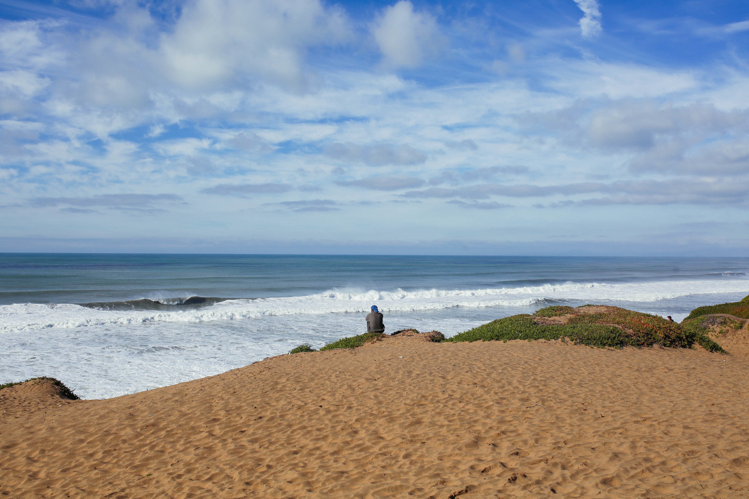 Person sitting on a sandy beach watching the ocean waves under a partly cloudy sky.