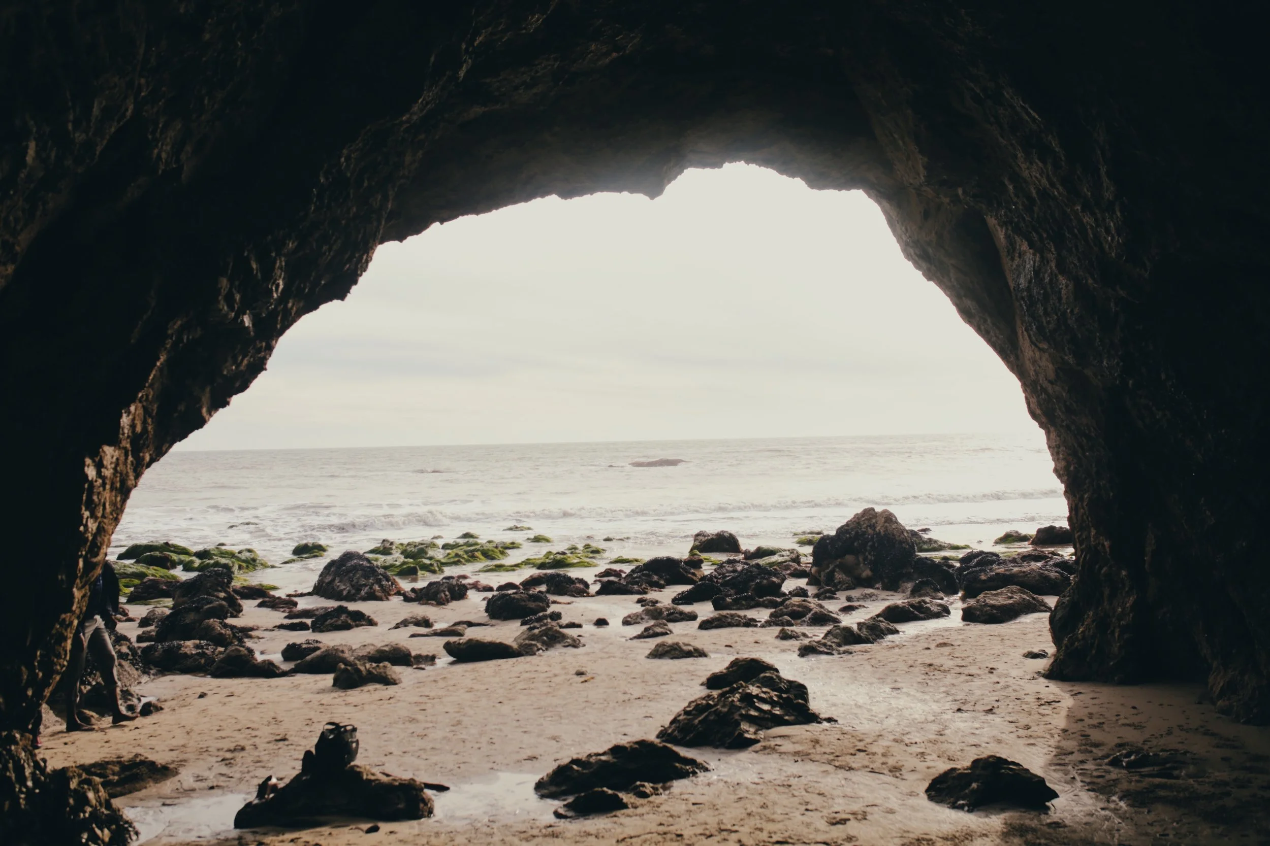 View from inside a rocky cave looking out to a sandy beach with rocks leading to the ocean and cloudy sky.