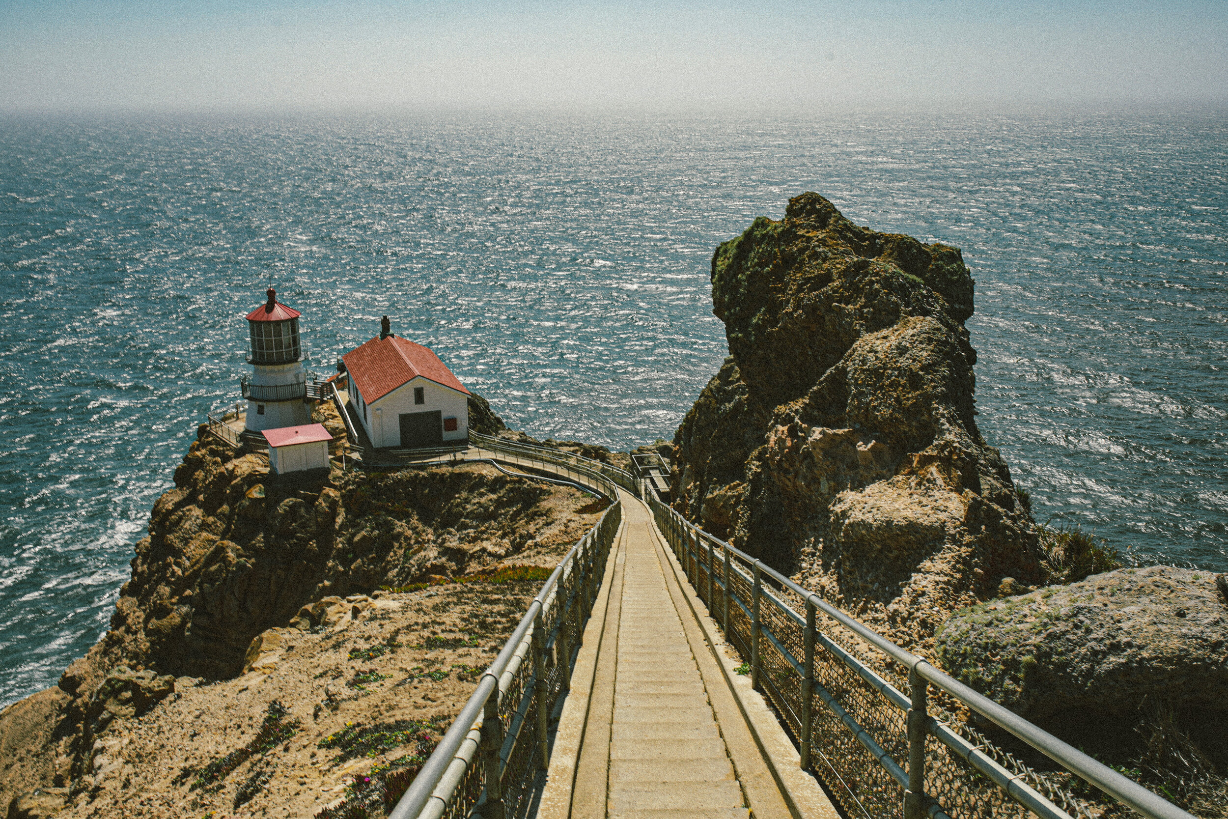 Pathway leading to a lighthouse and small house on a rocky cliff above the ocean with a large rock formation nearby.