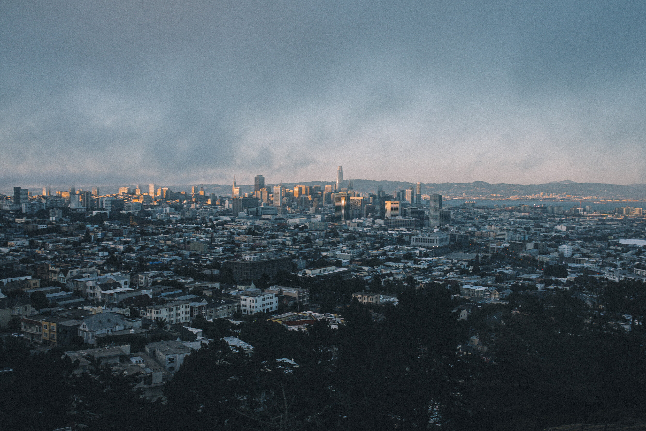 City skyline of San Francisco with tall buildings and the Bay in the background, under a cloudy sky.