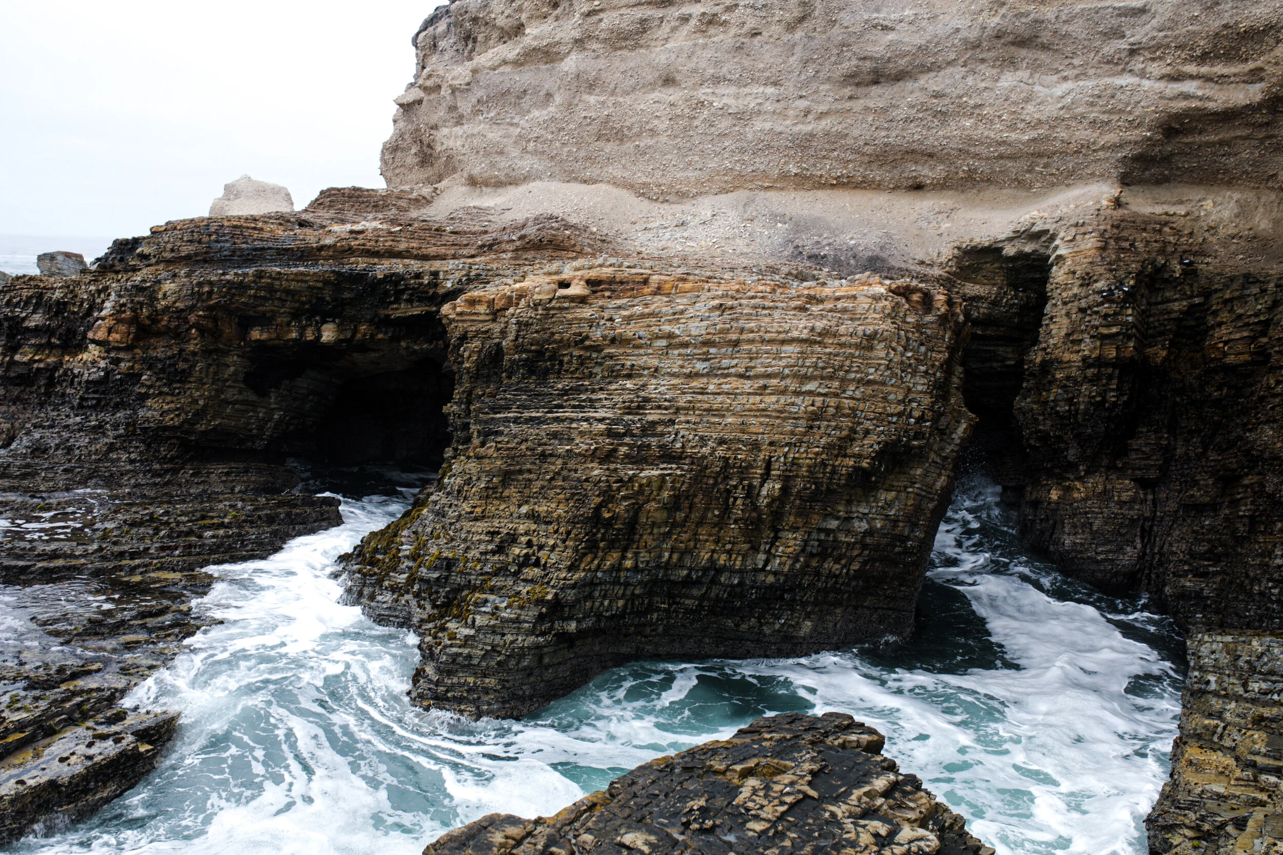 Coastal rock formations with a large cave and waves crashing against the rocks.