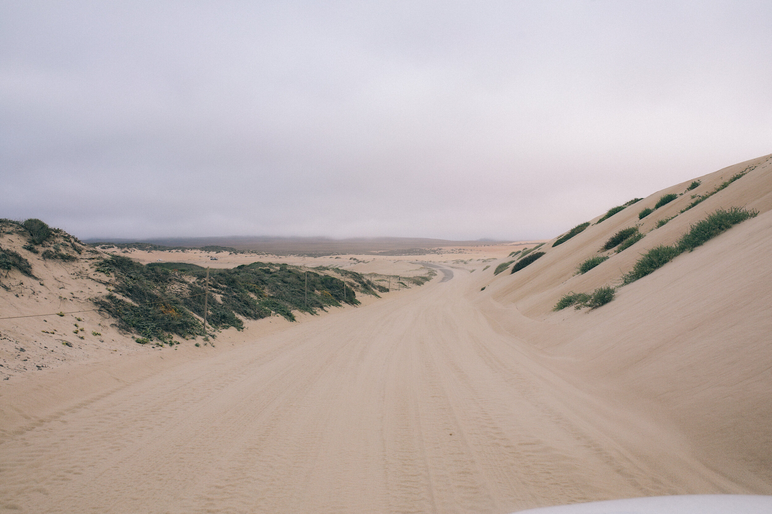 A sandy dirt road winding through a semi-arid landscape with hills and sparse vegetation under a cloudy sky.