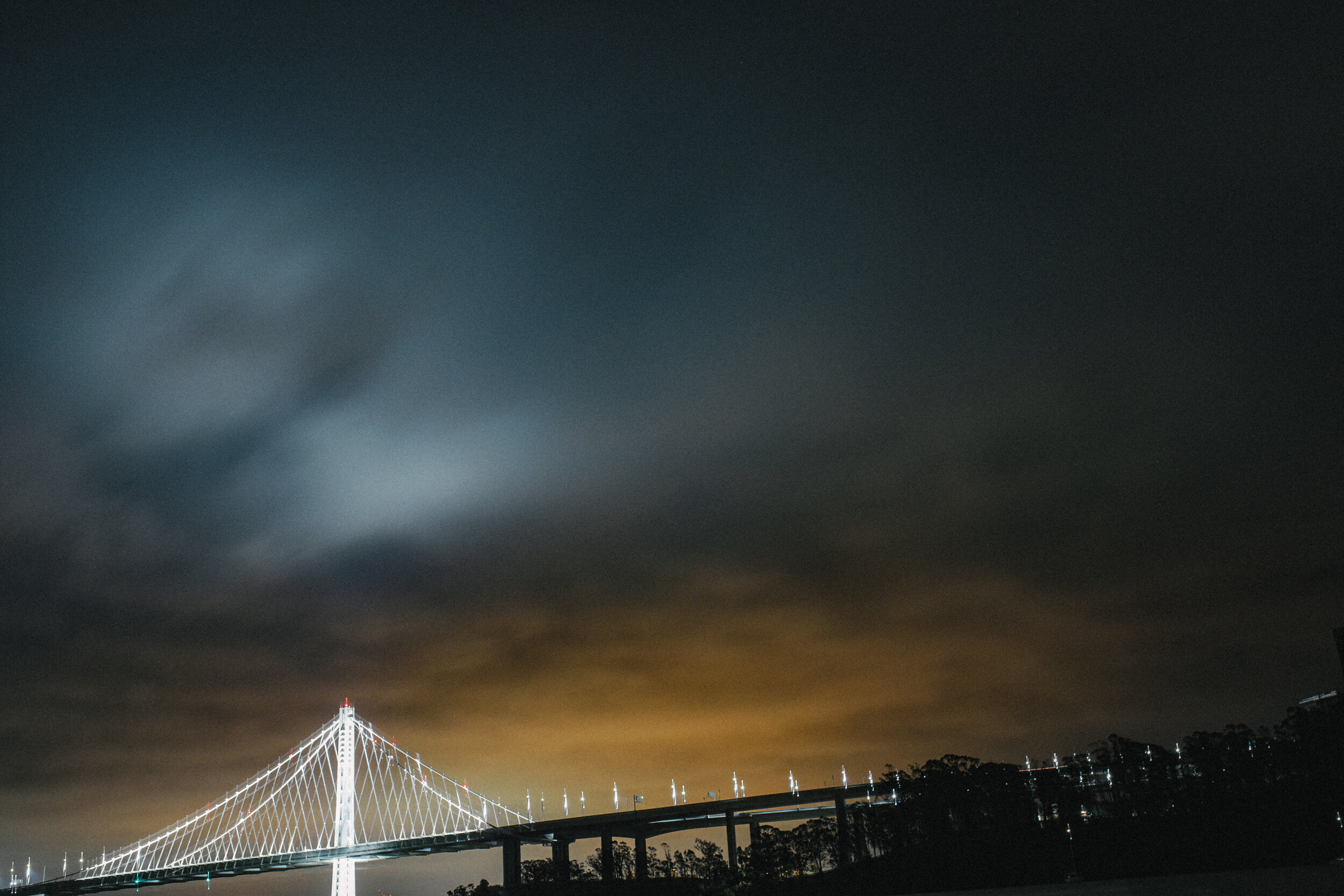 Nighttime photograph of a lit-up suspension bridge with a dark cloudy sky overhead and trees in the background.