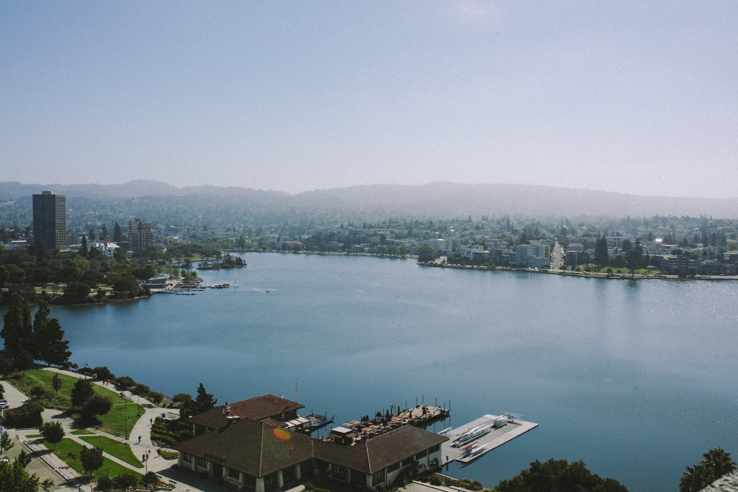 Aerial view of a cityscape with a large lake in the foreground, buildings along the shoreline, and hills in the background under a clear sky.