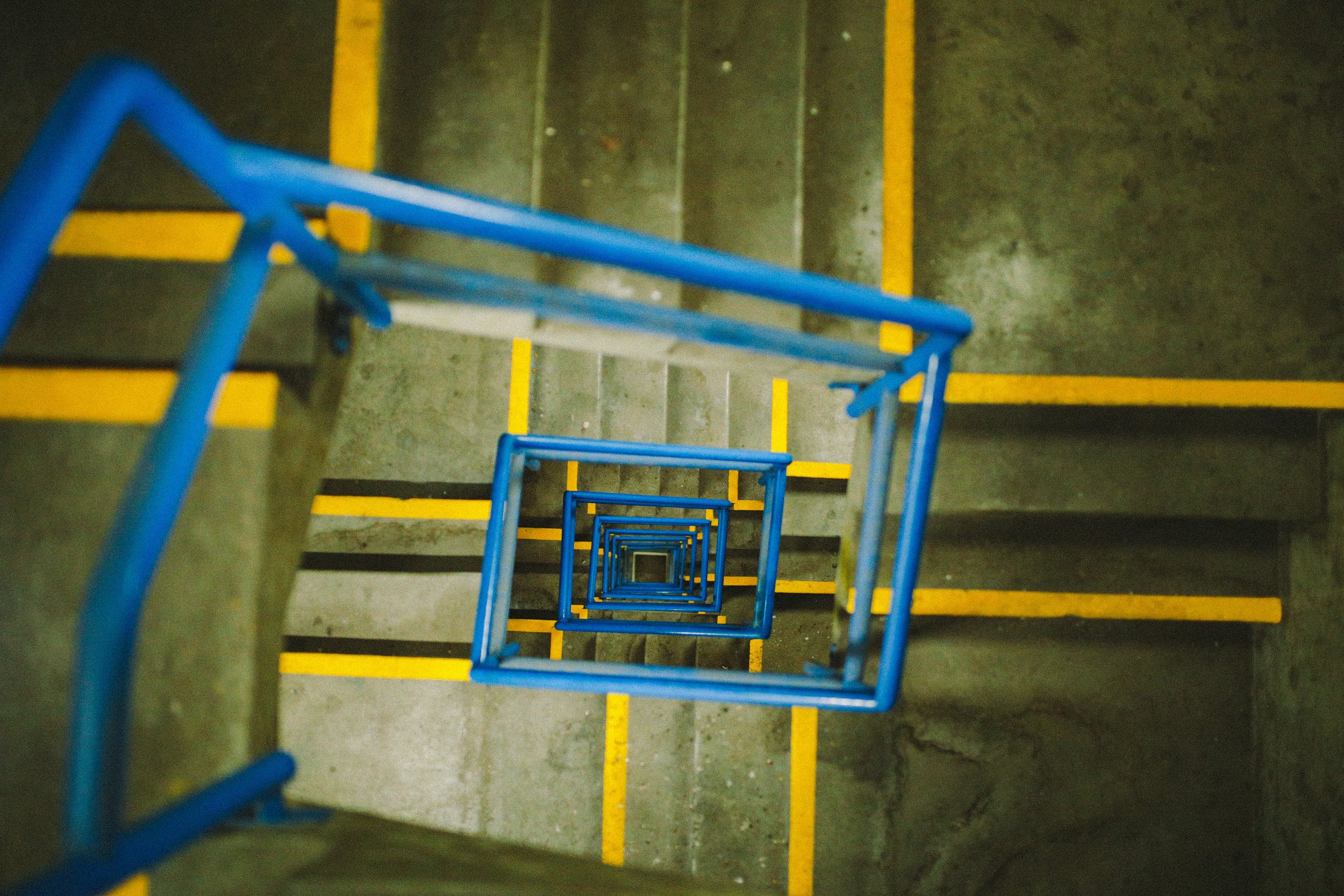 Looking down a stairwell with yellow safety markings on each step and a blue metal handrail, creating a repeating tunnel effect.