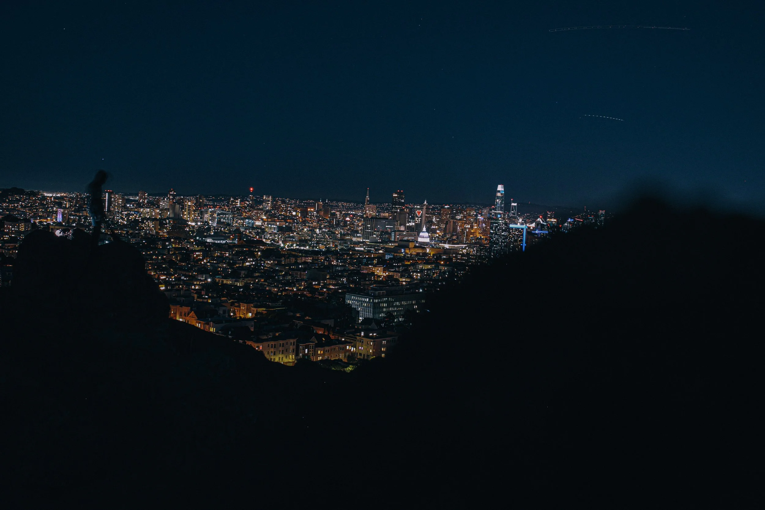 Nighttime cityscape view of San Francisco skyline with illuminated buildings, skyscrapers, and a clear sky with a few star trails.