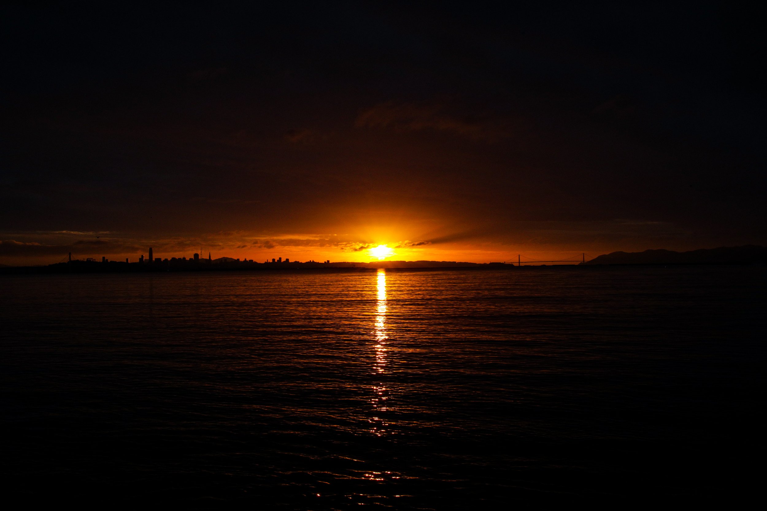 Sunset over the city skyline with reflections on the water and a bridge in the distance.