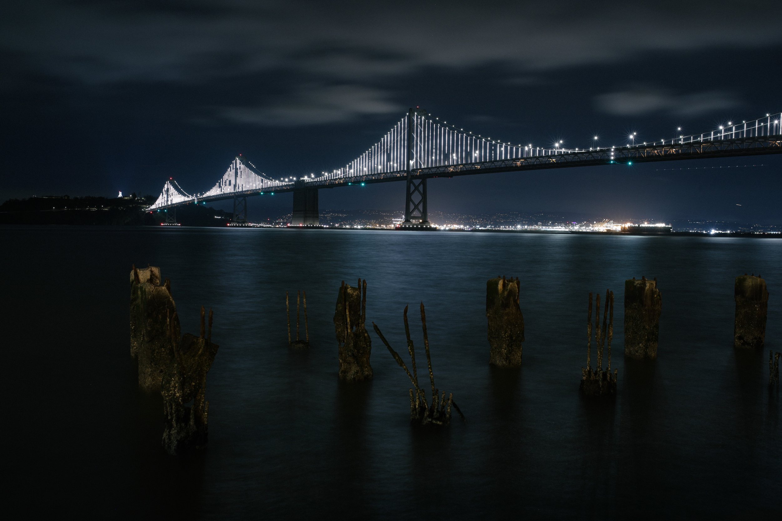 Night view of a lit suspension bridge over water, with old wooden posts and steel rebar remnants in the foreground.