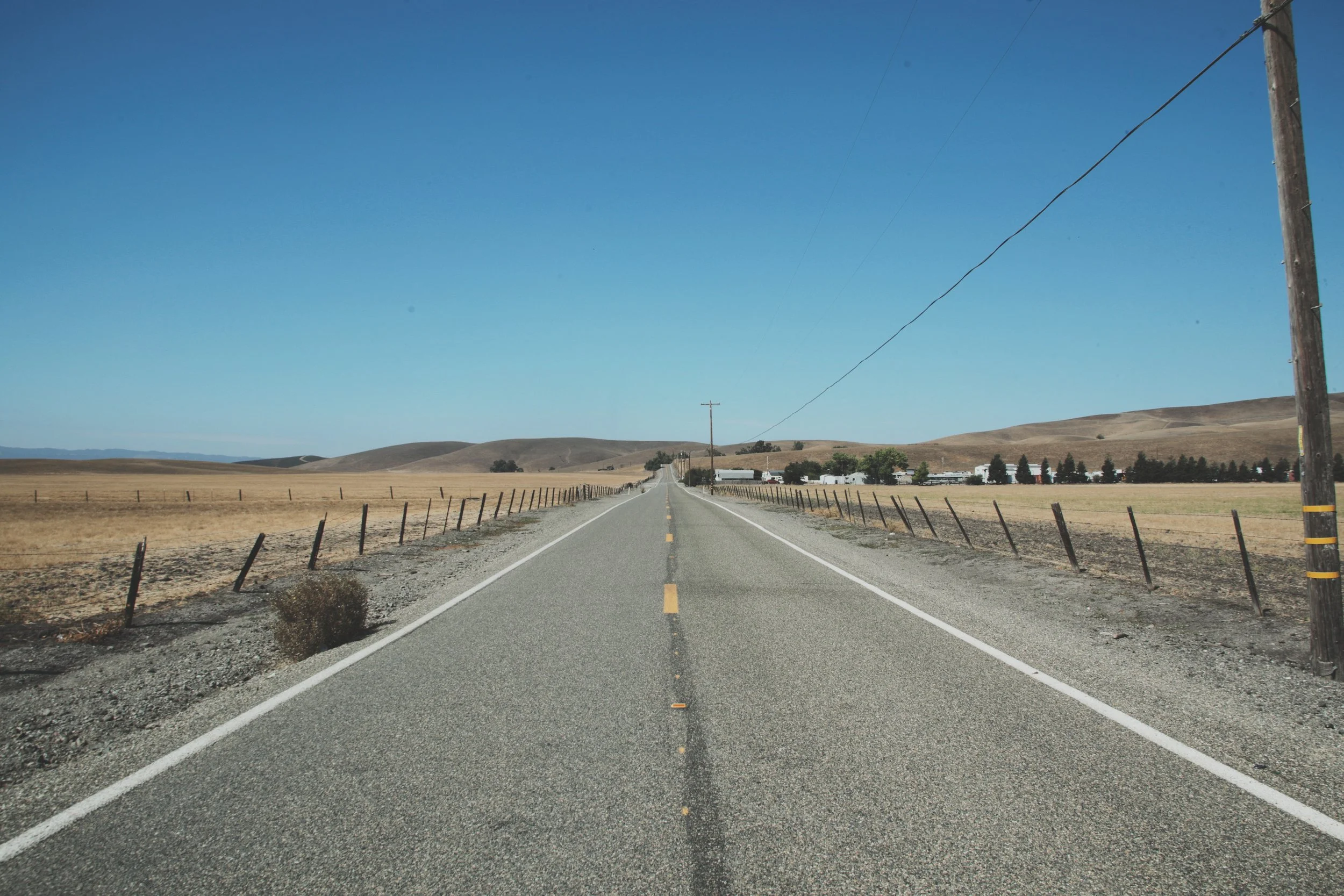 Long empty rural road with utility poles on the right and broken fence on both sides, leading towards distant hills under a clear blue sky.