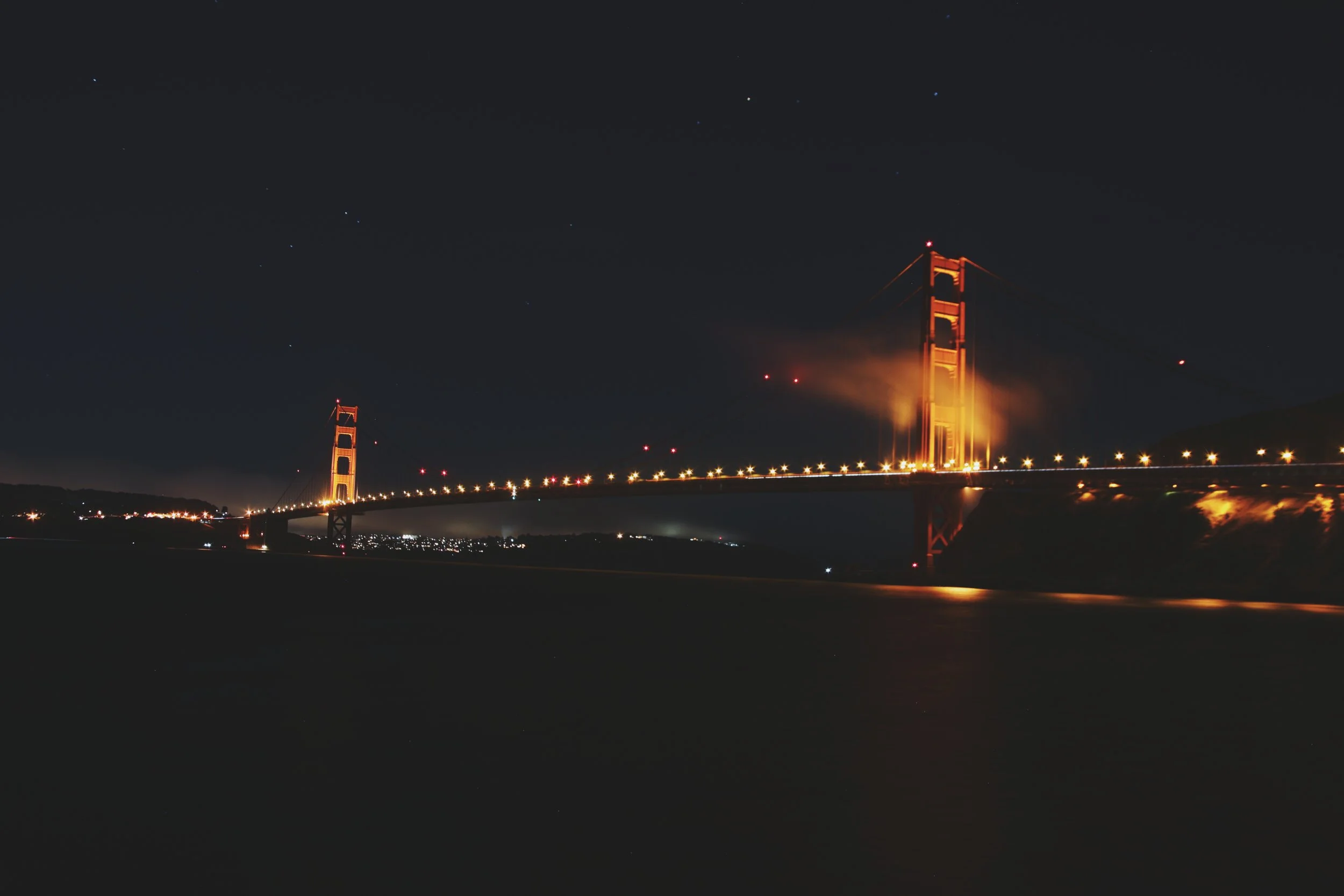Night view of the Golden Gate Bridge illuminated in orange lights, with fog and starry sky in the background.
