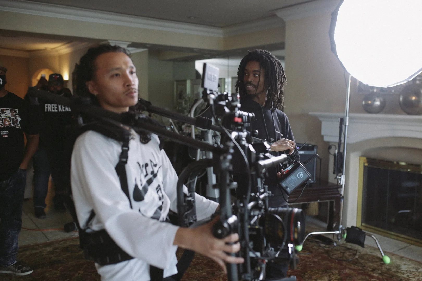 Two filmmakers on a set, one woman with camera equipment, another man observing, in a room with a large studio light and a fireplace.