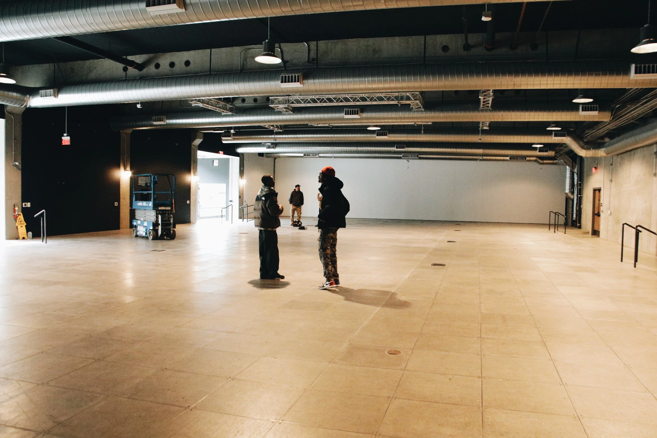 An empty indoor space with four people standing and talking. The space has a high ceiling with exposed ductwork, large open floor, and industrial design elements.