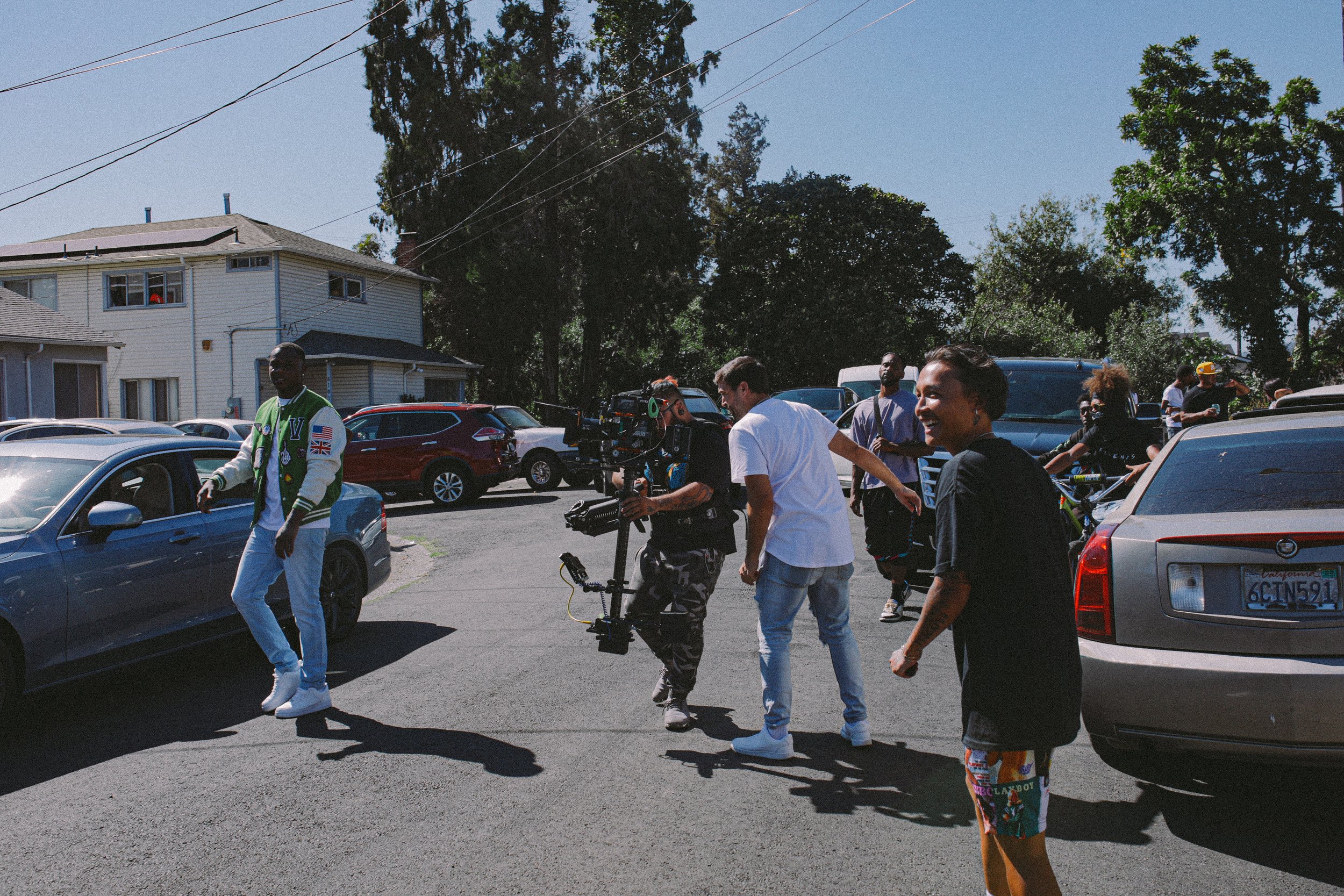 A film crew working on a street parking lot with multiple people and equipment, including a camera operator and crew members, some smiling and talking among parked cars and residential buildings in the background.