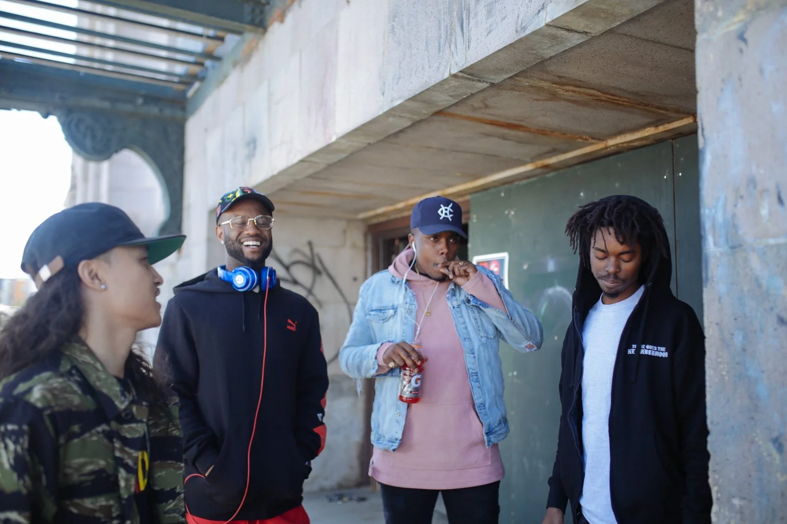 Four young adults standing together outdoors in front of a green wall and a grey exterior wall, engaging in conversation and smiling.