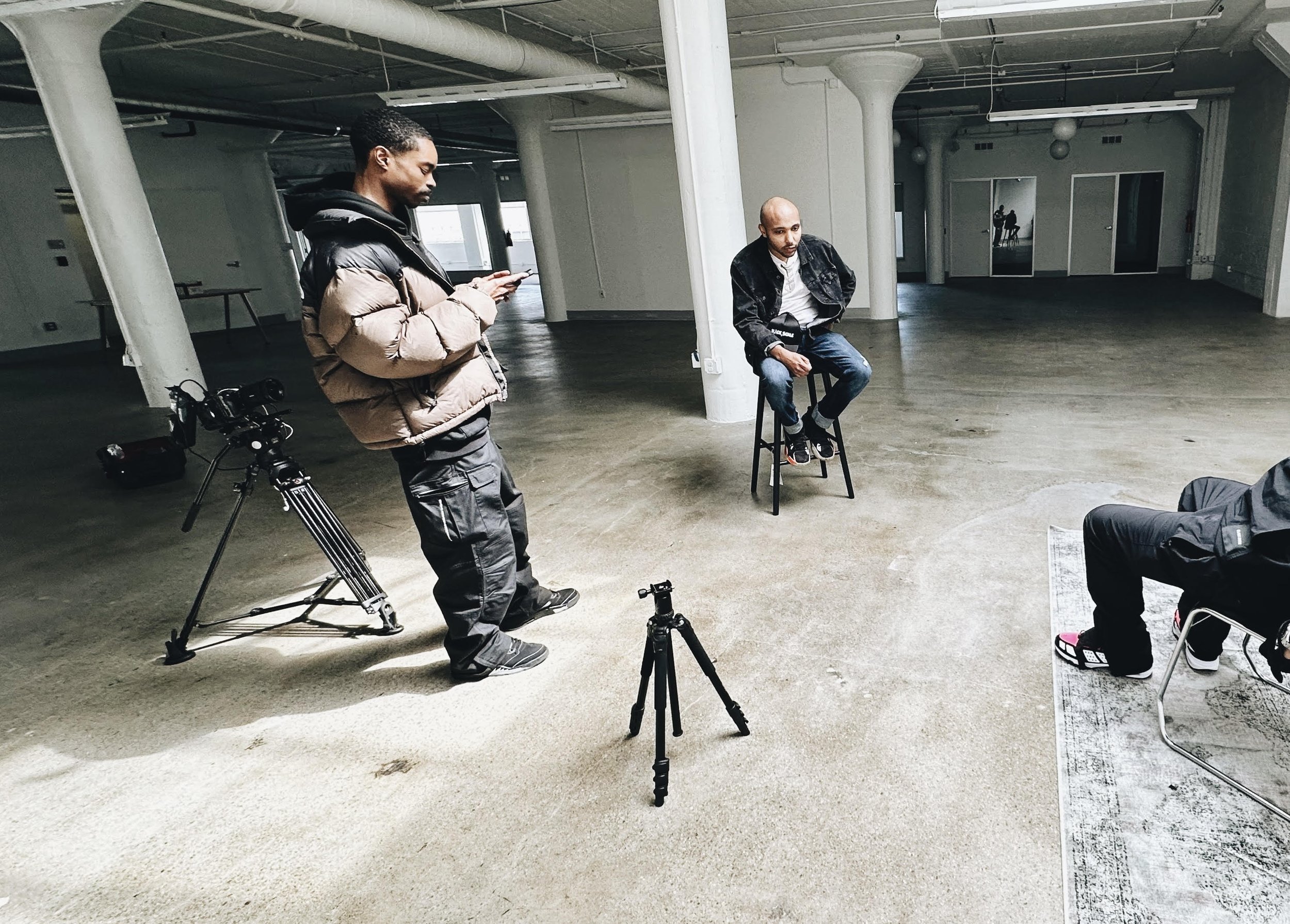 Three men in a spacious, industrial-style room engaged in a photo shoot. One man is sitting on a stool, the second man is standing and looking at his phone, and the third man is sitting on a chair. Photography equipment including tripods and a camera is present.
