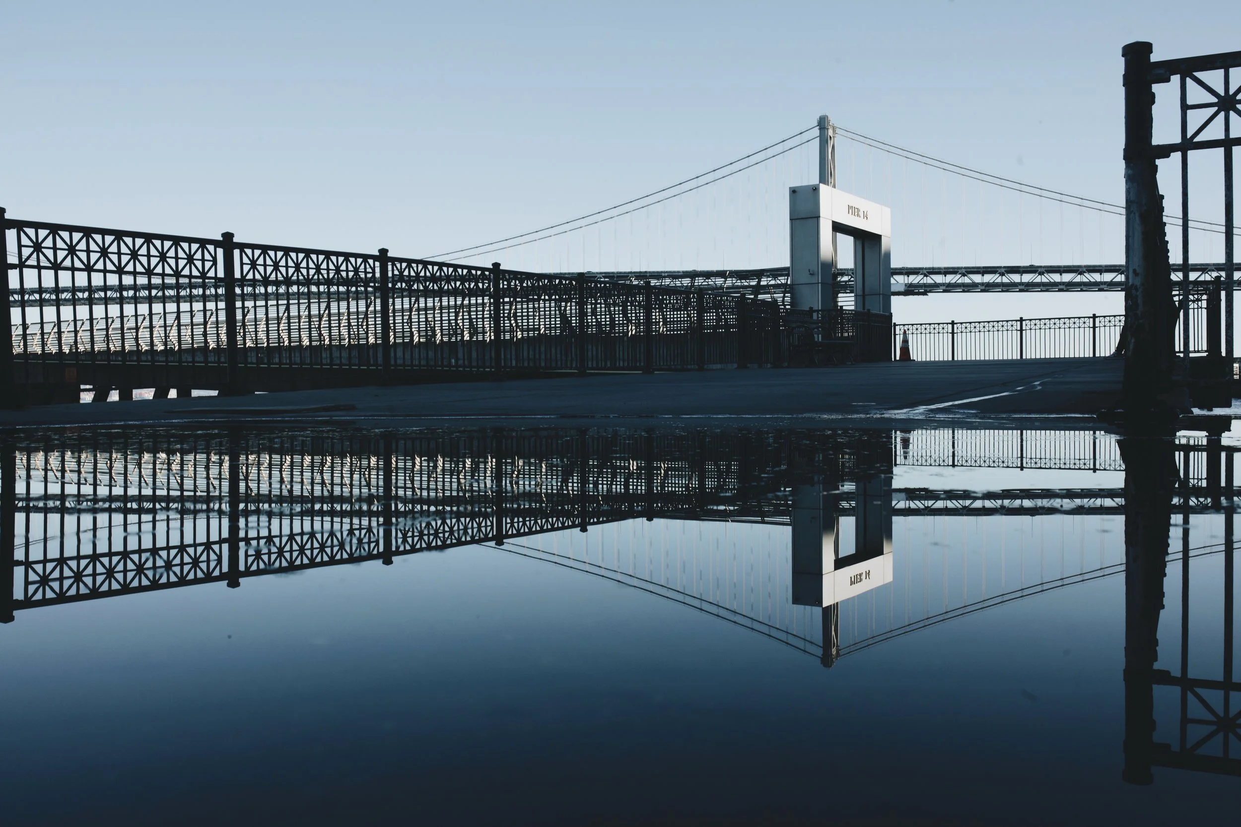 A bridge with a metal railing and a reflection in a large puddle of water on the ground, with a clear blue sky in the background.