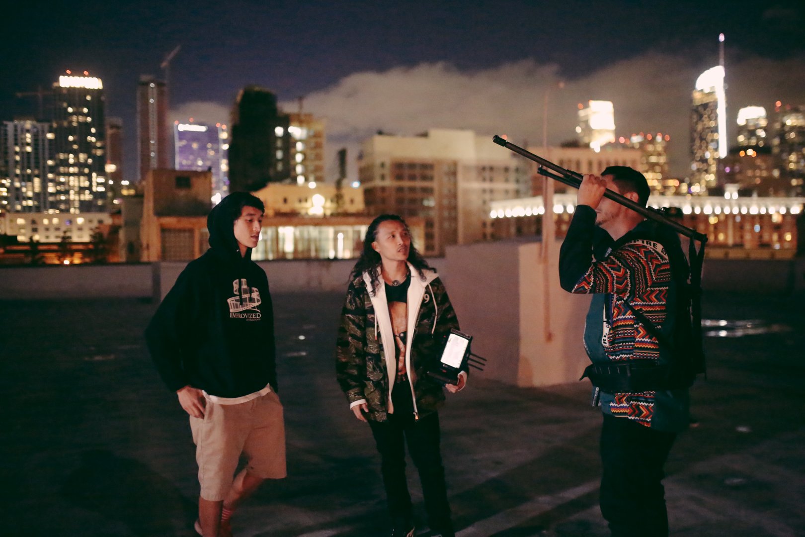 Three people on a rooftop at night in a city, one holding a long pole, another holding a device, with city lights and buildings in the background.