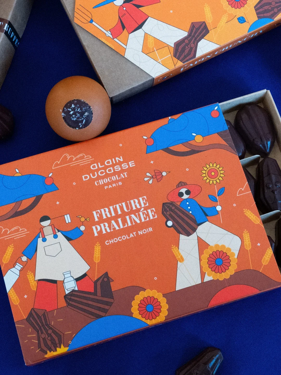 Boîte de chocolat noir Alain Ducasse avec illustration colorée de deux personnages, un homme avec un chapeau rouge et une femme en robe bleue, entourés de fleurs, de blé et d'autres éléments de la nature.