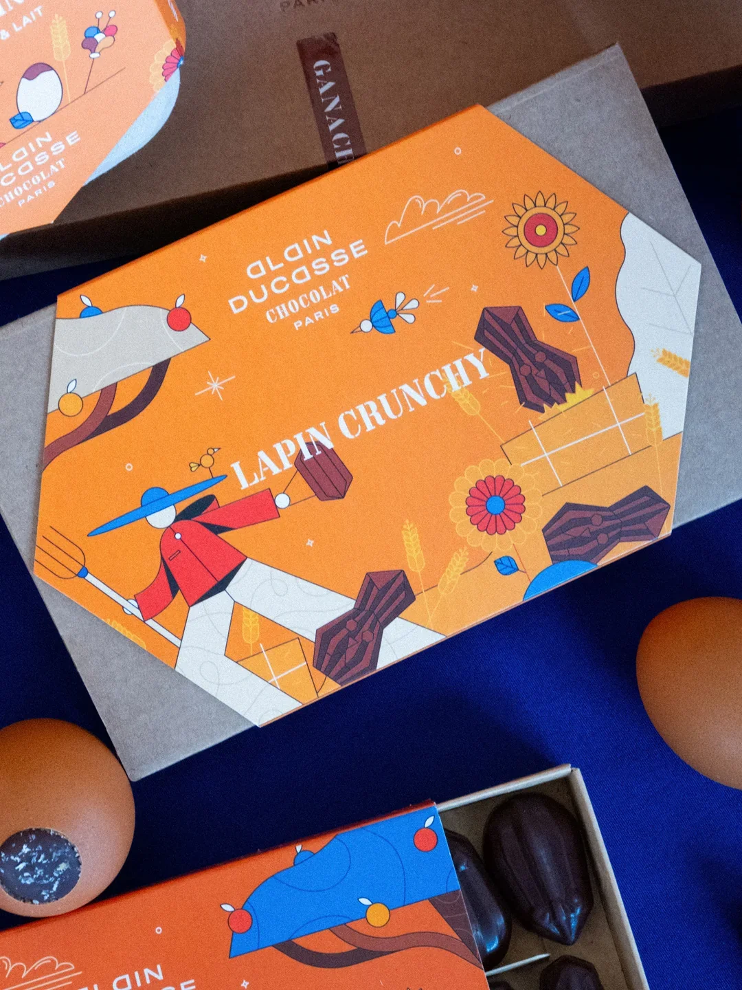 Boîte de chocolat Ducasse Paris avec un design coloré représentant un personnage avec un grand chapeau bleu, entouré de fleurs et de grains de blé, avec une ambiance printanière.