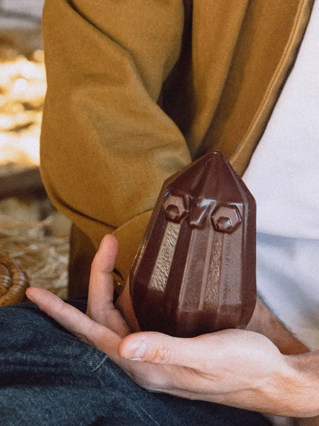 Une personne tenant un objet en forme de pyramide en chocolat, avec un vêtement marron et un fond beige.