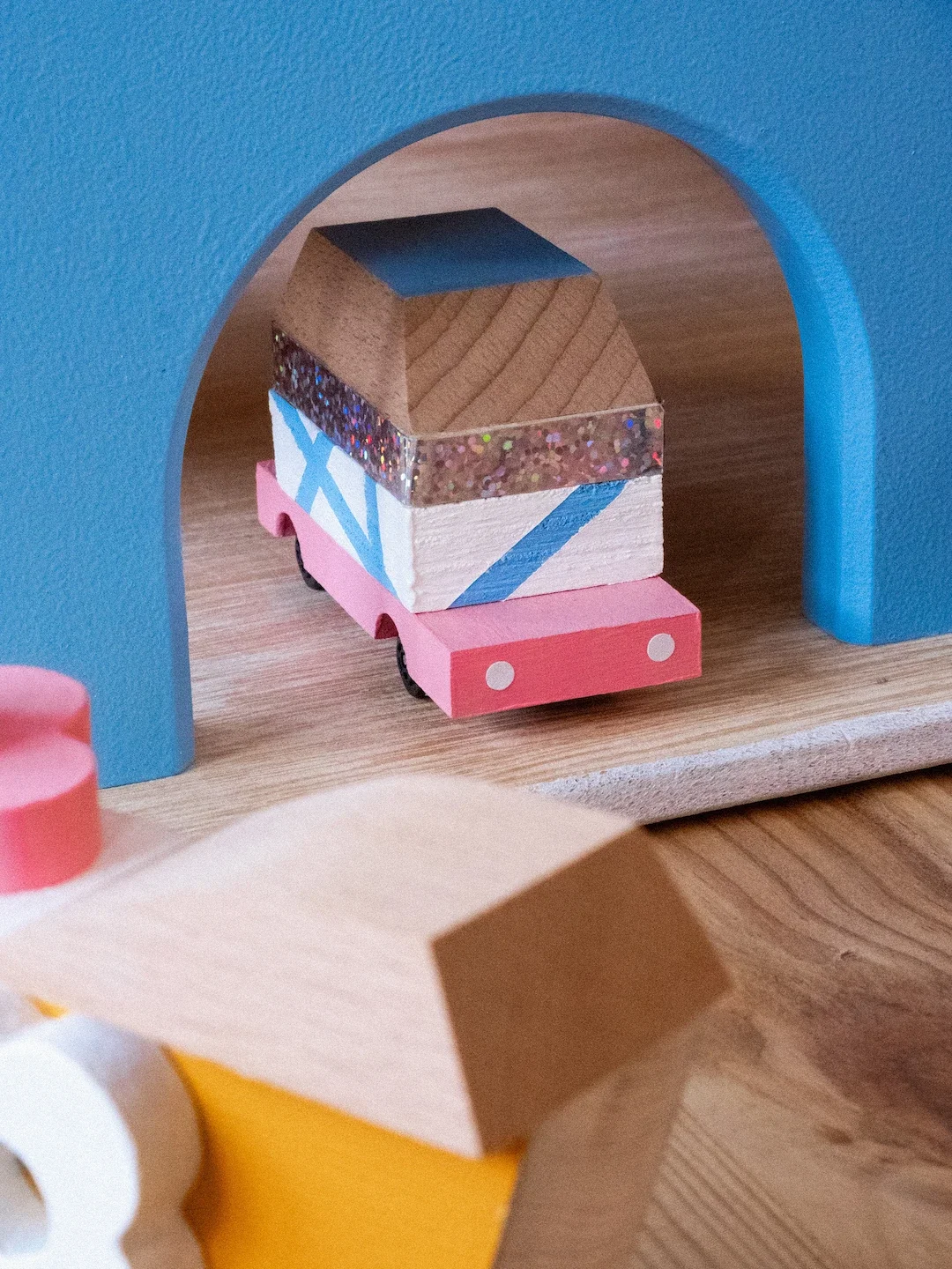 Une voiture en bois rose avec des cercles blancs sur le pare-chocs, passant sous un pont en bois peint en bleu.