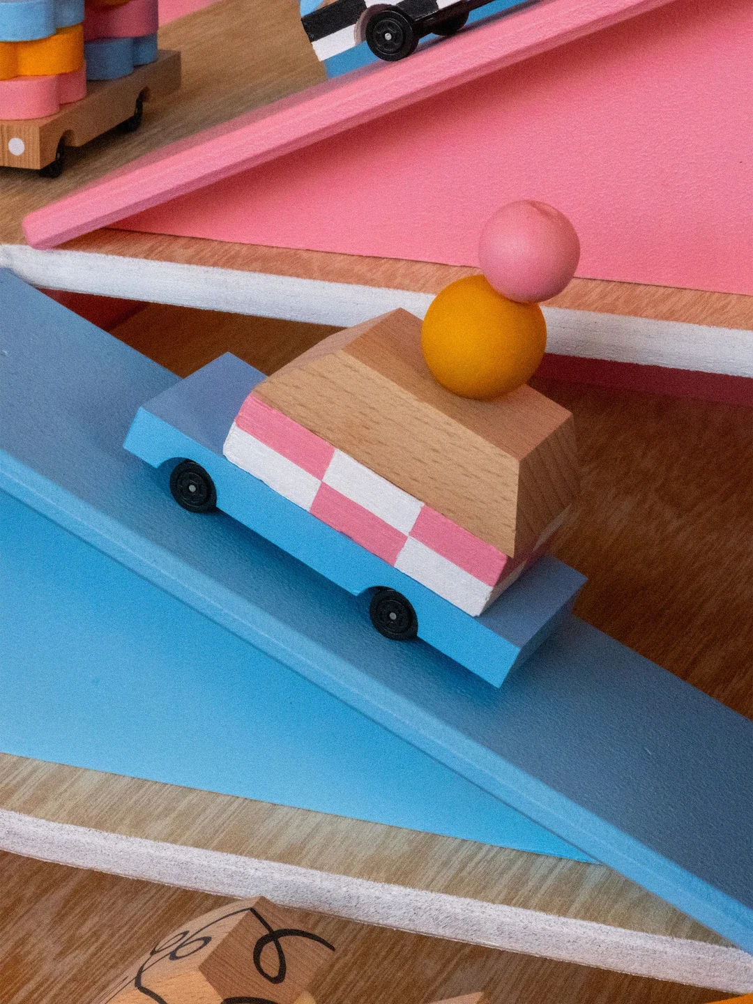 Une course de petites voitures en bois colorées avec des éléments en bois sculpté. La voiture est bleu avec un toit bleu clair, et porte deux boules en bois, une rose et une jaune.