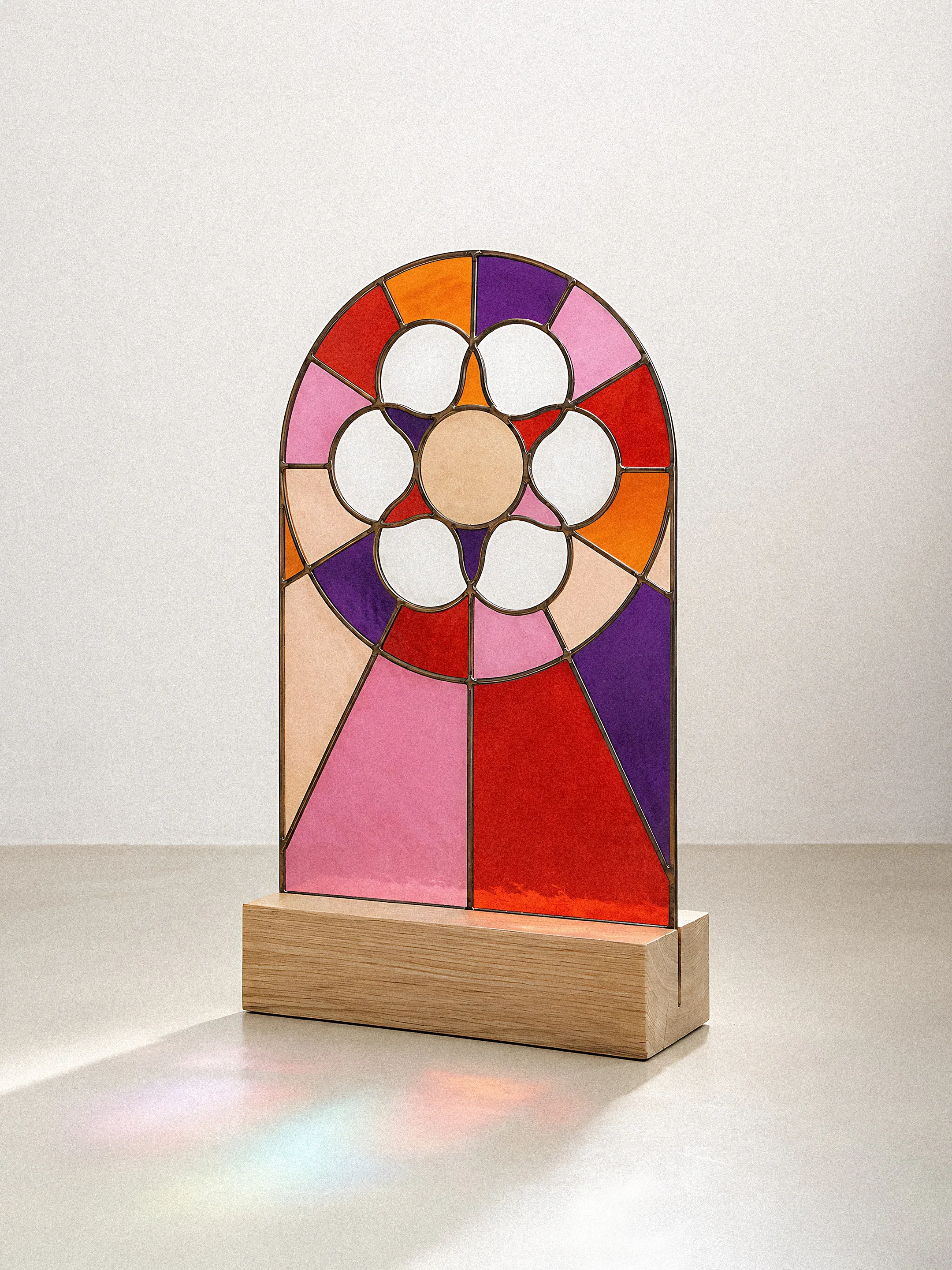 Une œuvre d'art moderne en vitrail coloré représentant un visage stylisé avec des formes géométriques, placée sur une base en bois, contre un fond neutre.