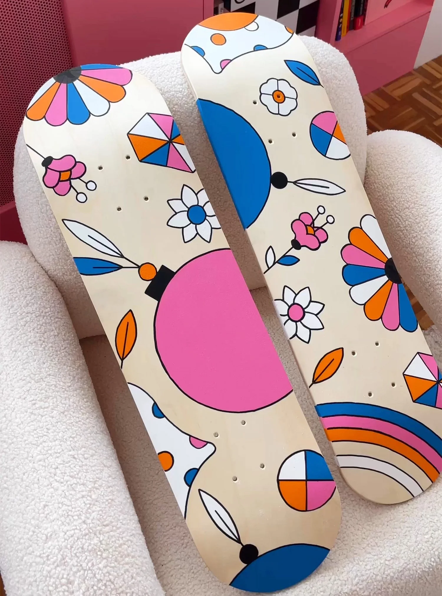 Deux planches de skateboard avec un design coloré de fleurs, de cercles et d'éléments géométriques sur un fauteuil en tissu blanc.