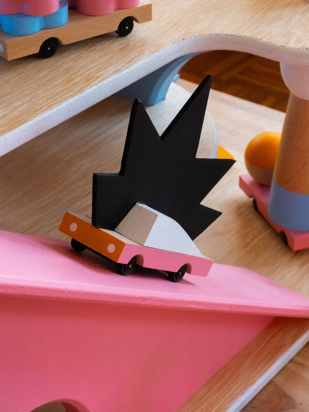 Décor en bois peint avec une silhouette de manga ou de super héros stylisée en noir, sur une plateforme rose avec roulettes, entouré d'autres éléments colorés en bois sur une surface en bois.
