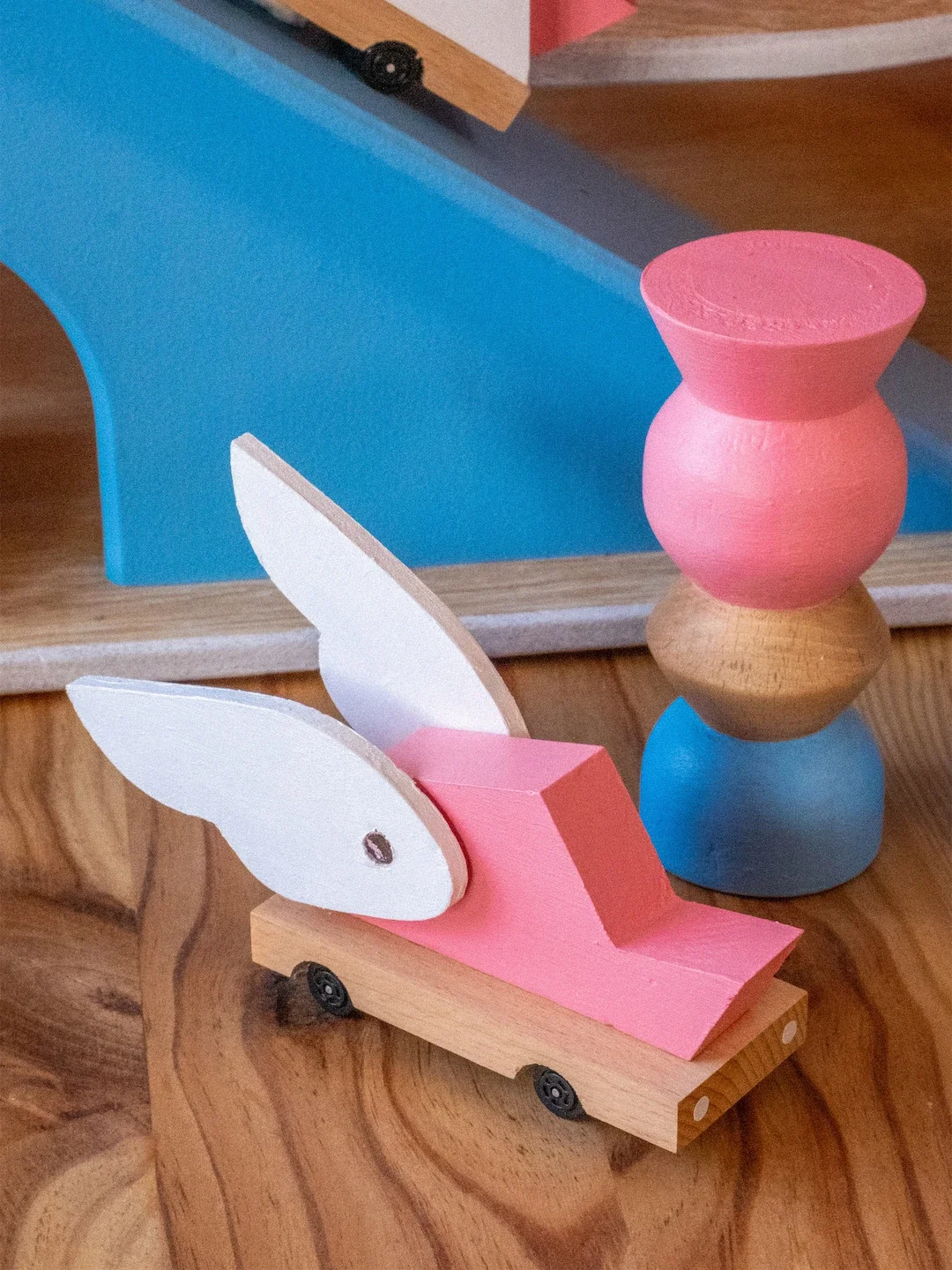 Figurine en bois d'un lapin avec ailes blanches, vieilli en rose, avec une base en bois, à côté d'une pile de trois formes en bois peintes en rose, beige et bleu, sur un meuble en bois.
