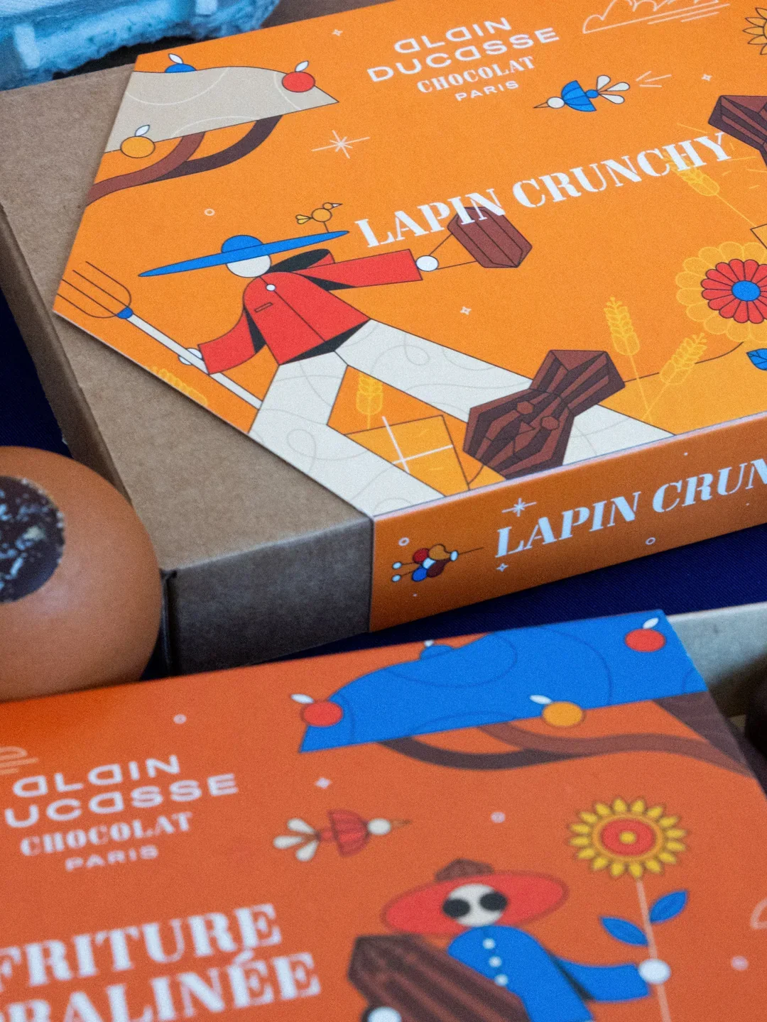 Emballages de chocolats de la marque Alain Ducasse, featuring des illustrations stylisées de lapins, poussant des chariots avec des barres de chocolat, sur un fond orange avec des motifs floraux et agricoles.