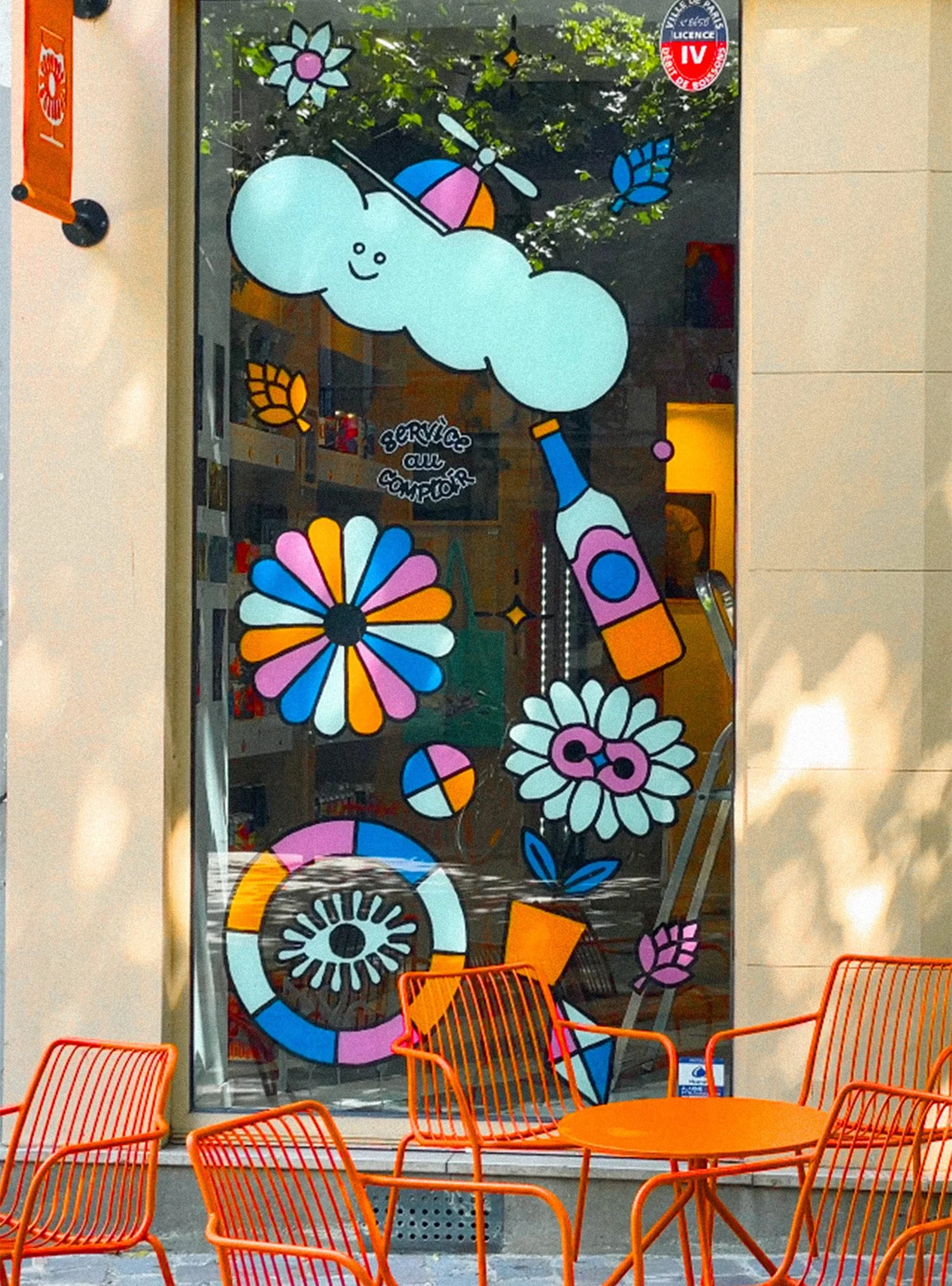 Fenêtre décorée avec des dessins colorés de fleurs, de nuages, d'une bouteille, d'un papillon et d'autres formes géométriques, avec des chaises orange et une table en avant.