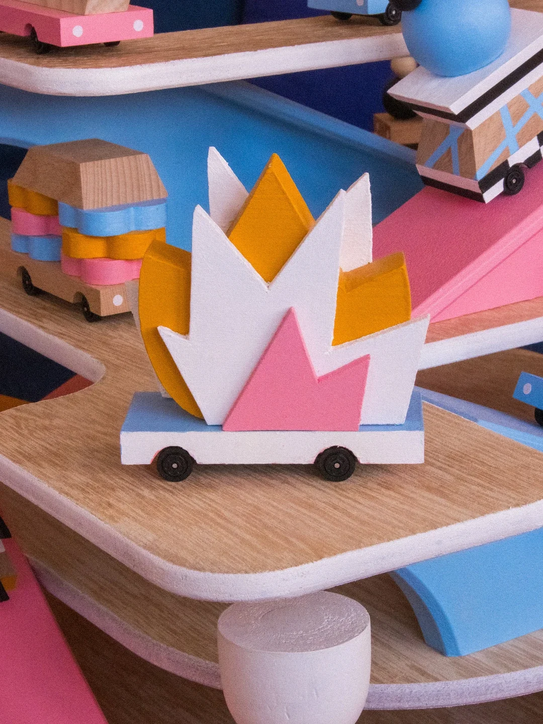 Une décoration de jouets en bois, représentant une flamme colorée avec des couleurs pastel comme le blanc, le jaune, le rose et le bleu, placée sur un chariot en bois avec des roues noires.