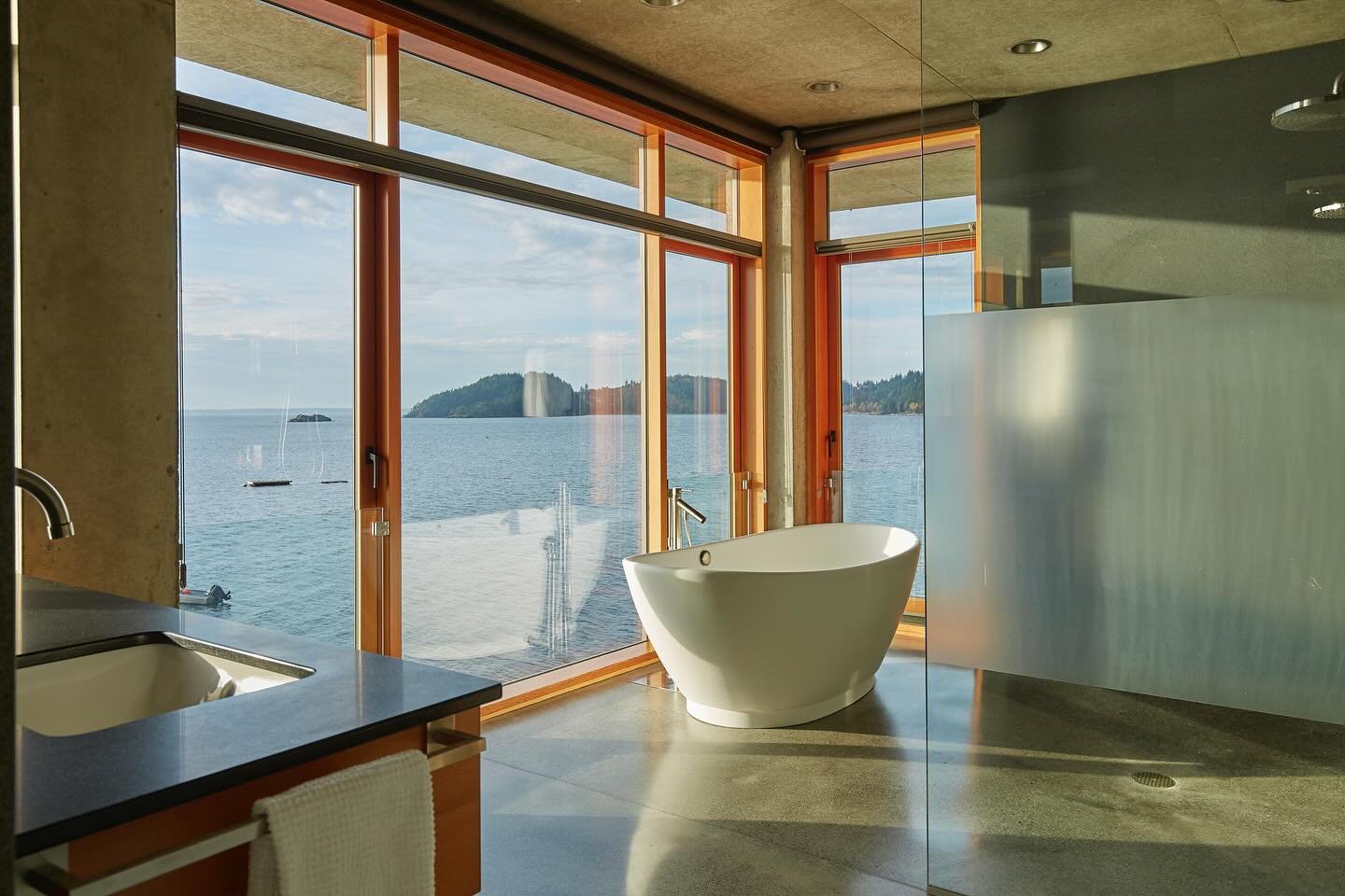 At the edge of Bellingham Bay, our #MCT_BayHouse Primary Suite feels as if it&rsquo;s floating above the water. This sense of levity balances the mass and weight of the all-concrete structure. A sculptural soaking tub anchors a glass-wrapped corner, 