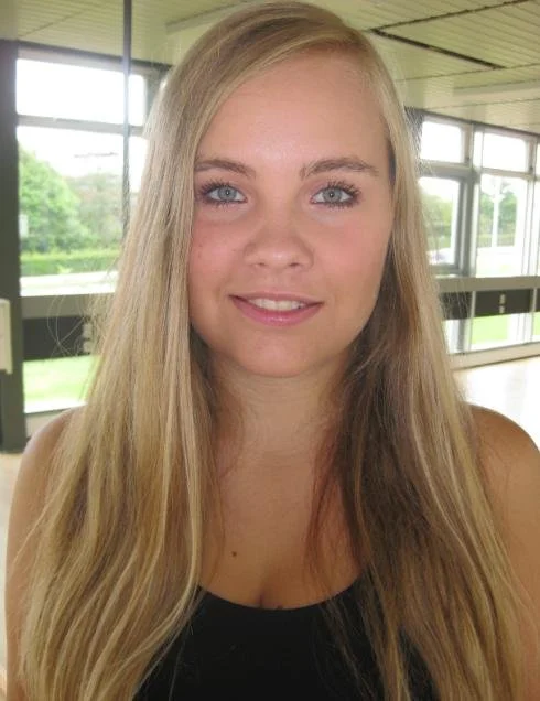 En ung kvinde med langt blondt hår, blå øjne og et smil, inde i et moderne bygning med store vinduer og grønne omgivelser udenfor. Danseskole i Aarhus