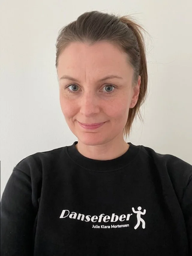 Kvinde med lyst hår i en hestehale, iført sort t-shirt med teksten »Dansefeber Julie Klara Mortensen« og en danserfigur. Danseskole i Aarhus
